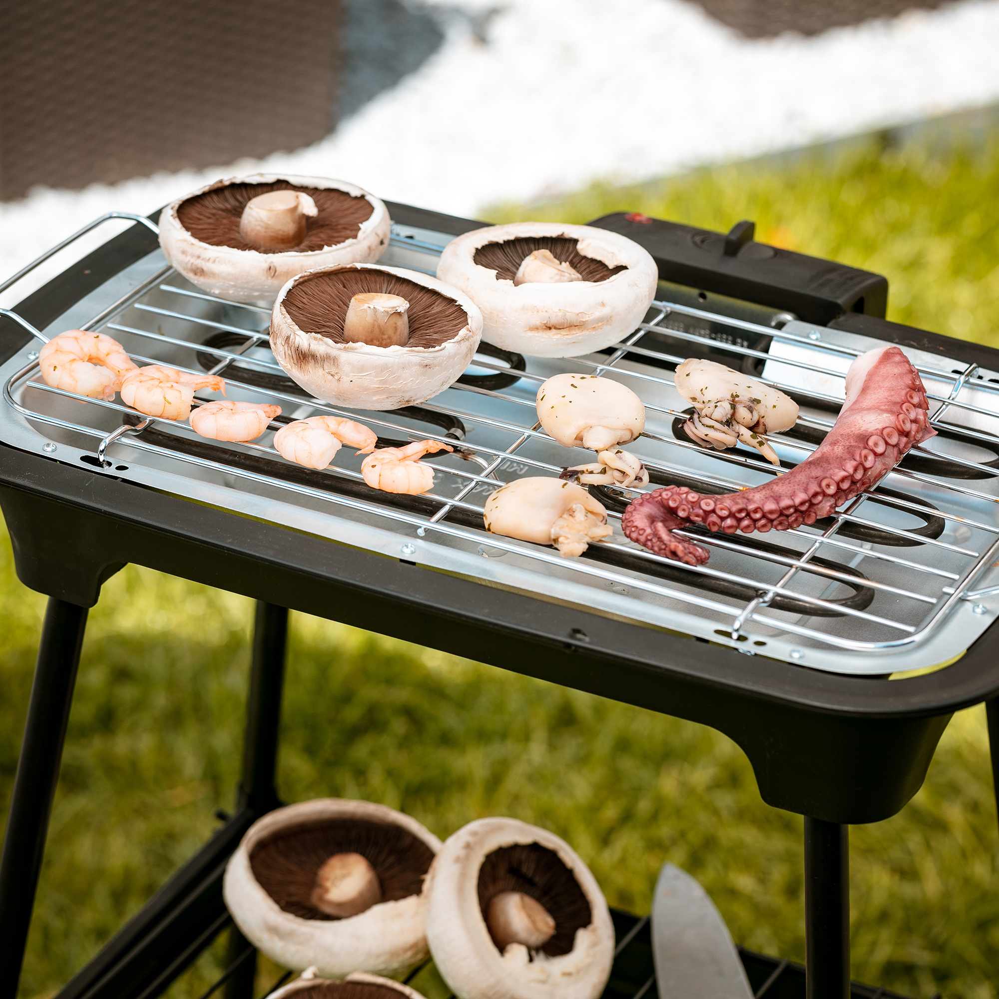 Grill mit Pilzen, Garnelen, Oktopus und anderen Lebensmitteln auf grünem Rasen.