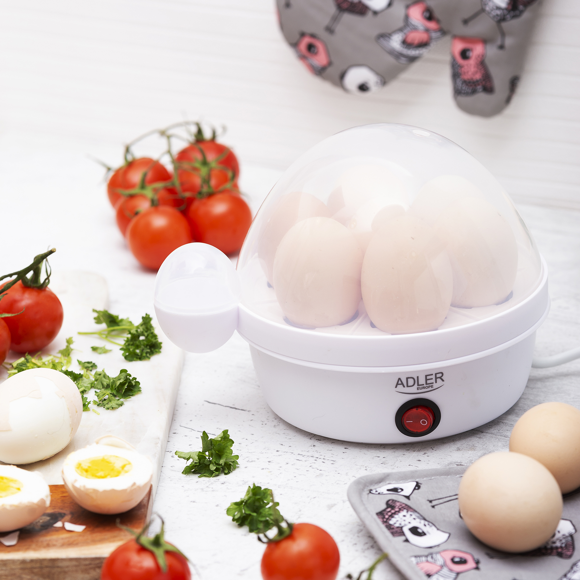 Hervidor de huevos blanco con huevos y tomates, sobre superficie blanca con tela y verduras.