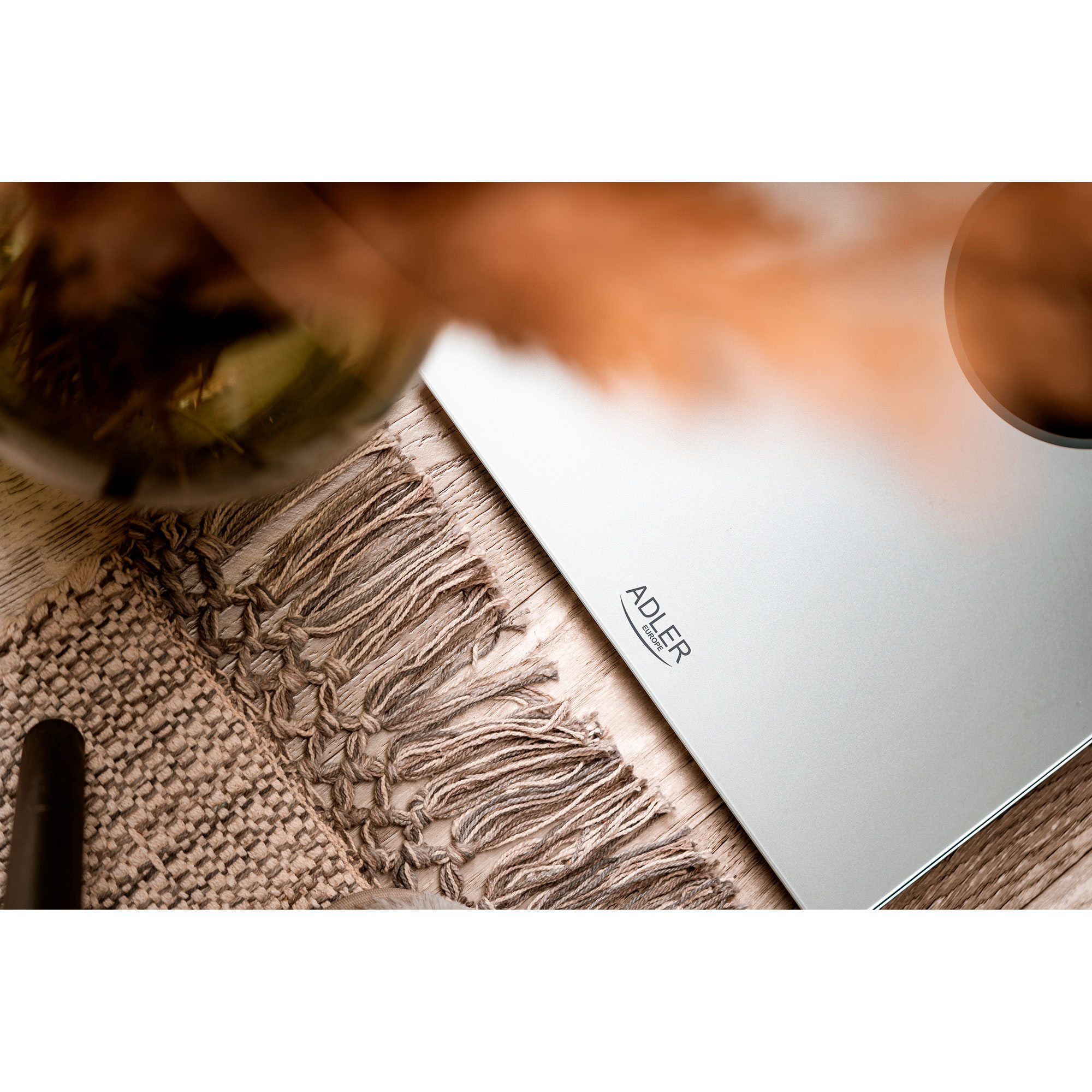 Srebrna waga z logo 'ADLER' na beżowym dywanie z elementami dekoracyjnymi.