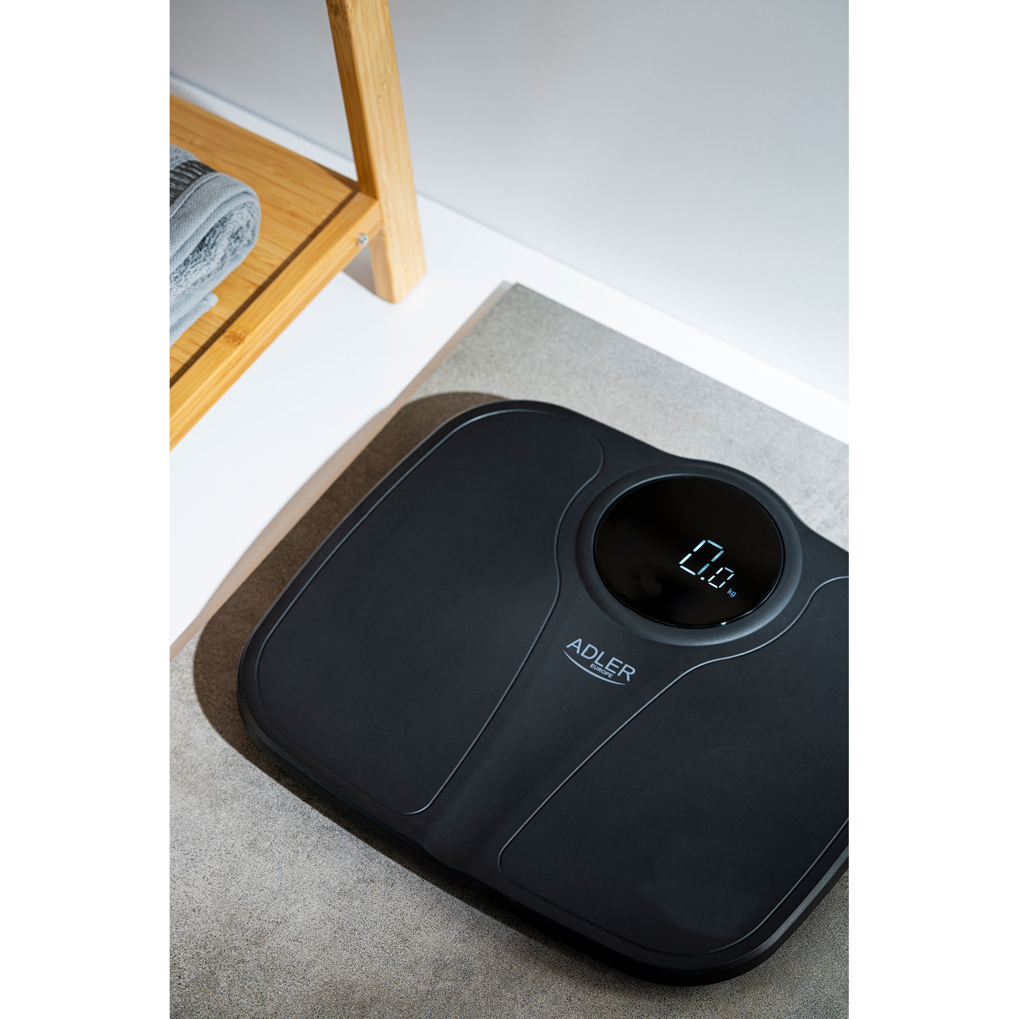 Eine schwarze digitale Badezimmerwaage zeigt 0,0 kg auf einer grauen Matte, mit einem Holzregal im Hintergrund.
