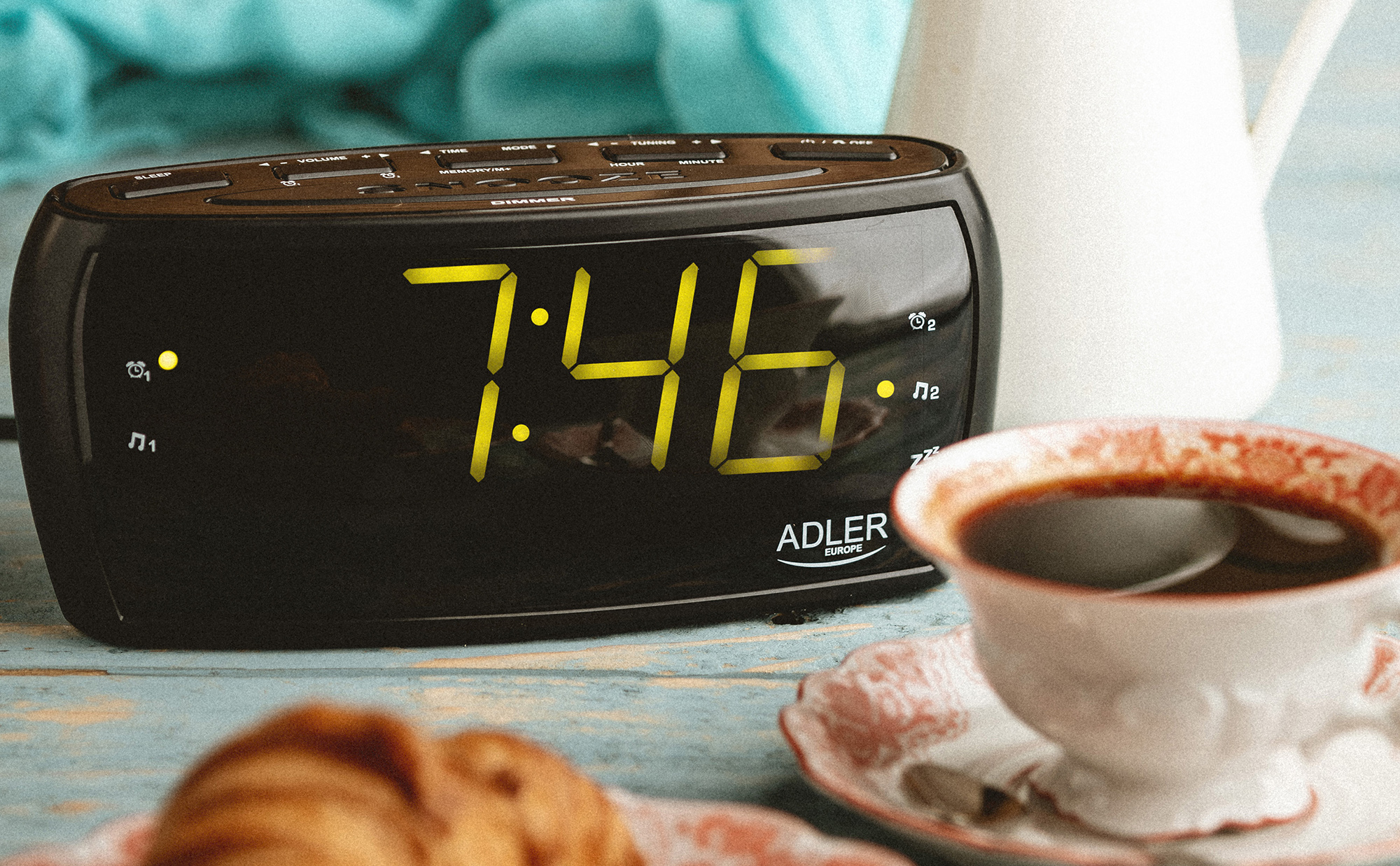 Despertador digital negro que muestra 7:46. Taza de café y plato visibles, escena matutina.