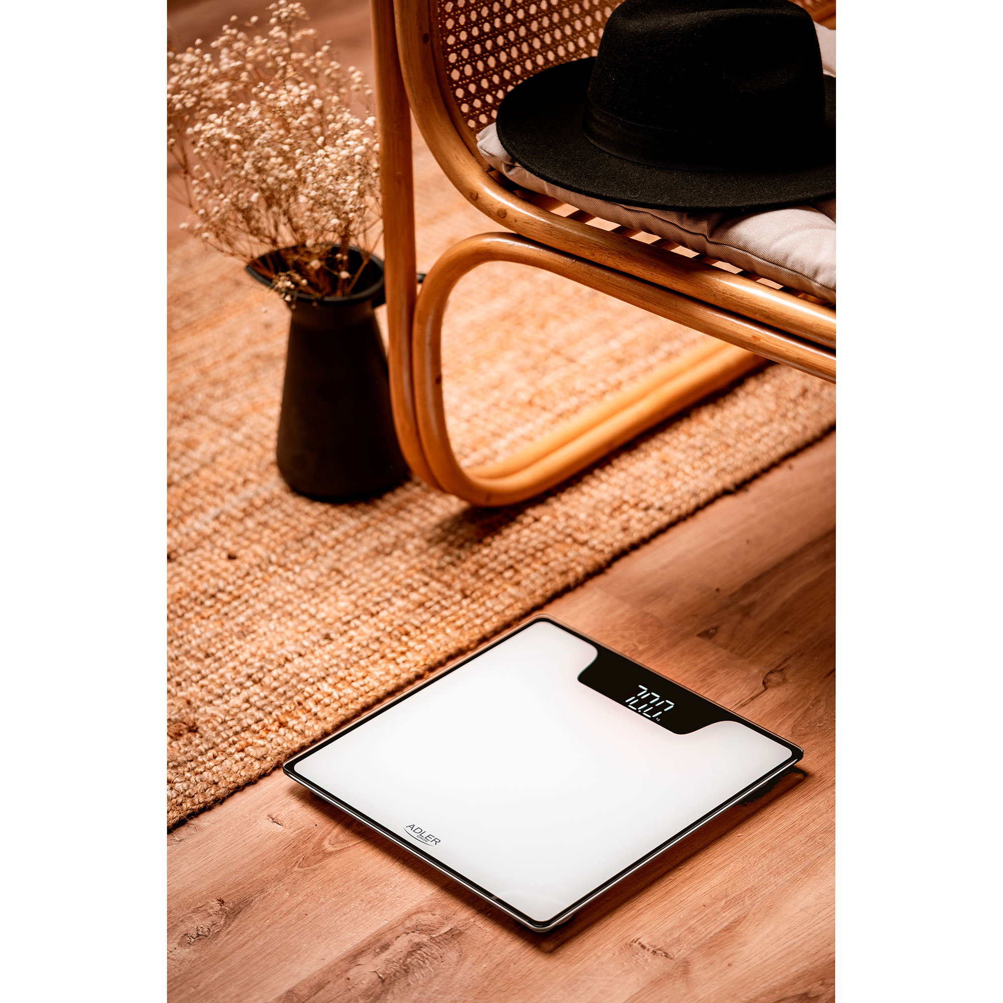 Waga cyfrowa na drewnianej podłodze obok dywanu, z kapeluszem i suszonymi kwiatami w tle.