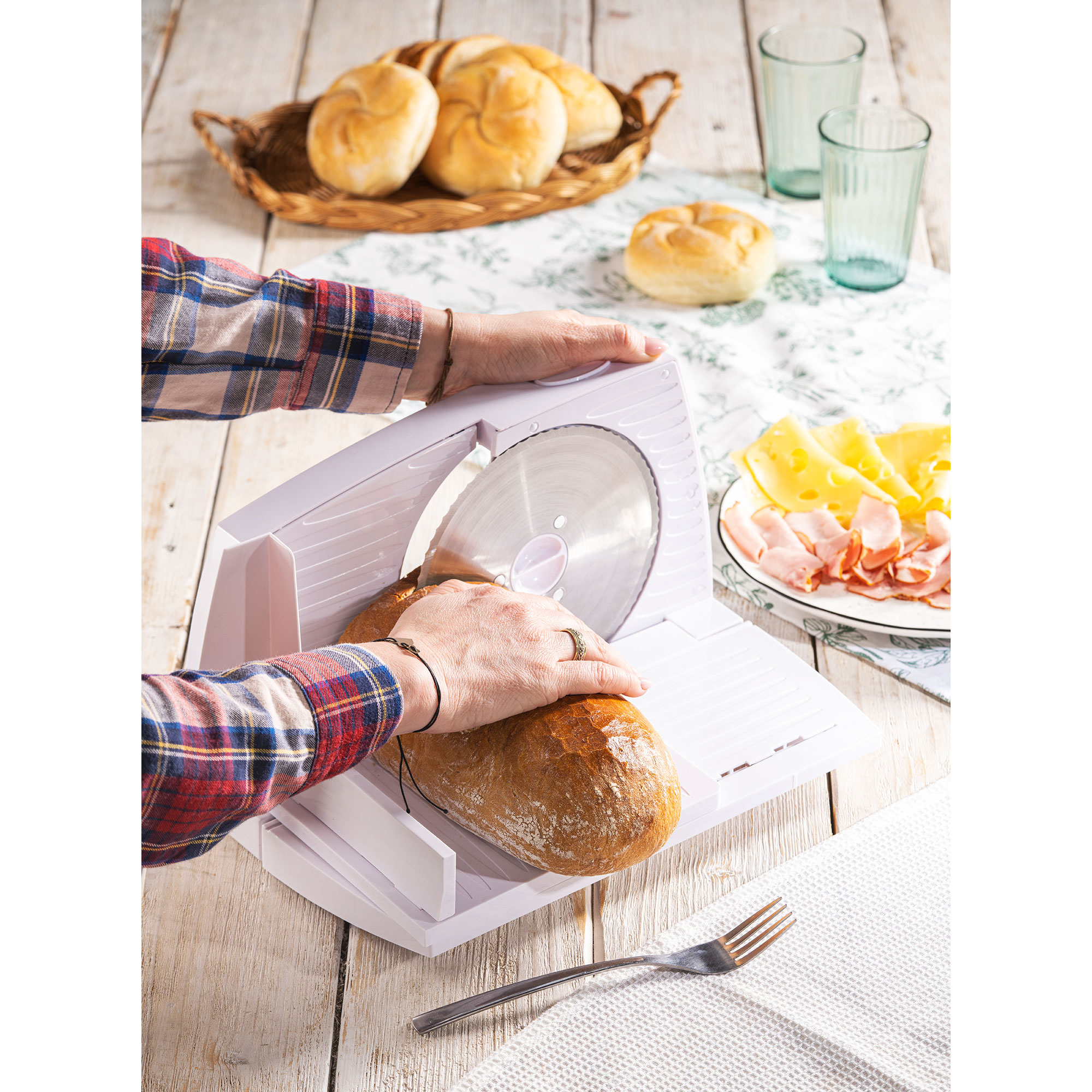 Osoba kroi chleb na białej krajalnicy z serem i szynką w tle.