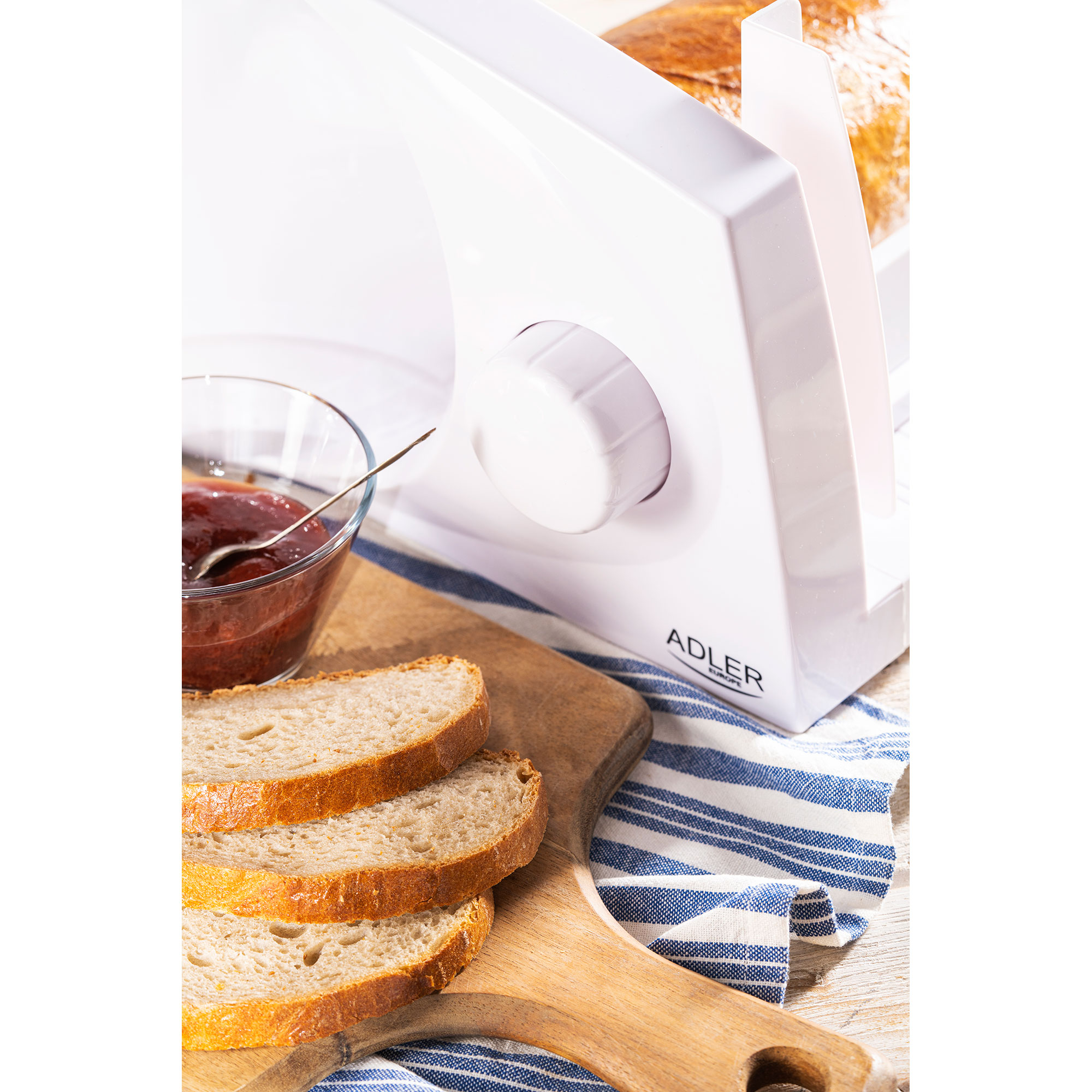 Biała krajalnica do chleba z bochenkiem chleba, kromkami i miską dżemu.