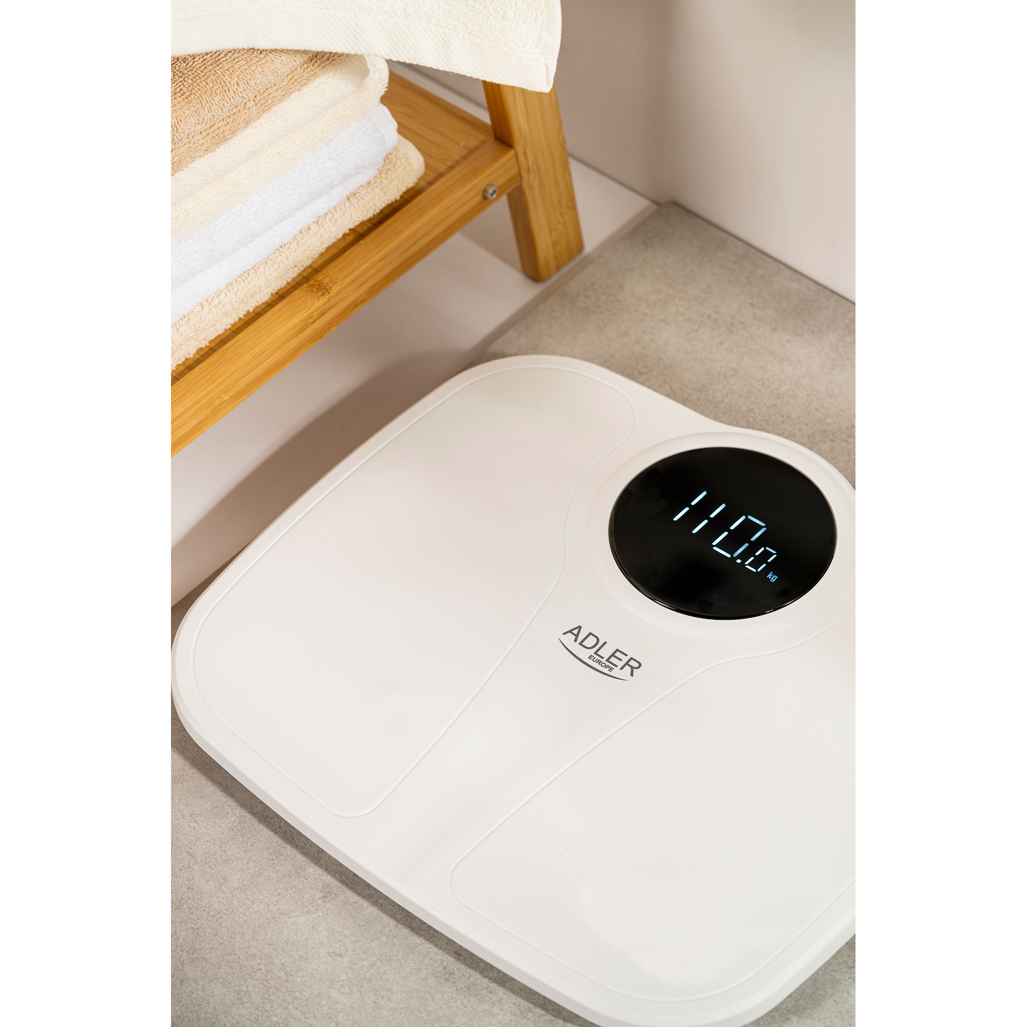 Biała waga cyfrowa pokazująca 110,0 kg, w pobliżu drewnianej półki z ręcznikami.