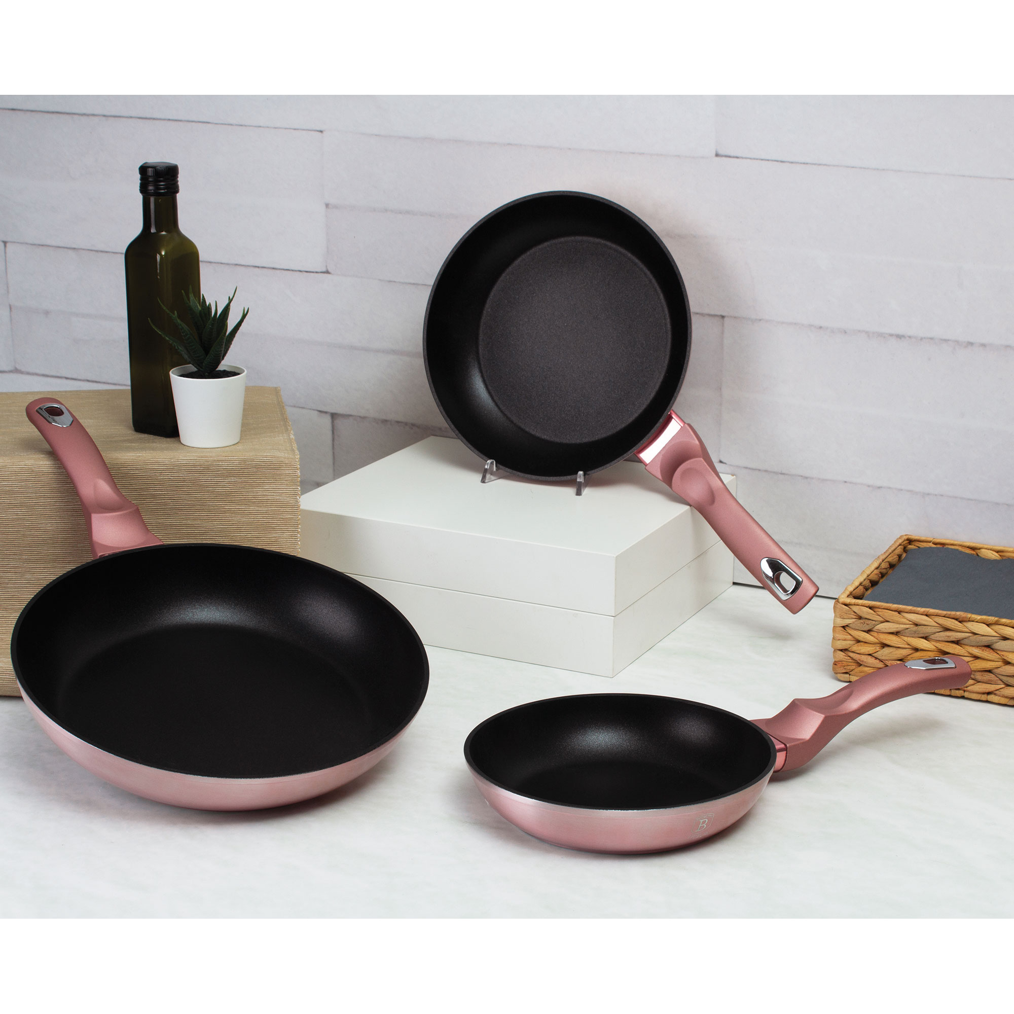 Tres sartenes de color oro rosa con interiores negros, una sobre un soporte. Fondo blanco y con diseño.