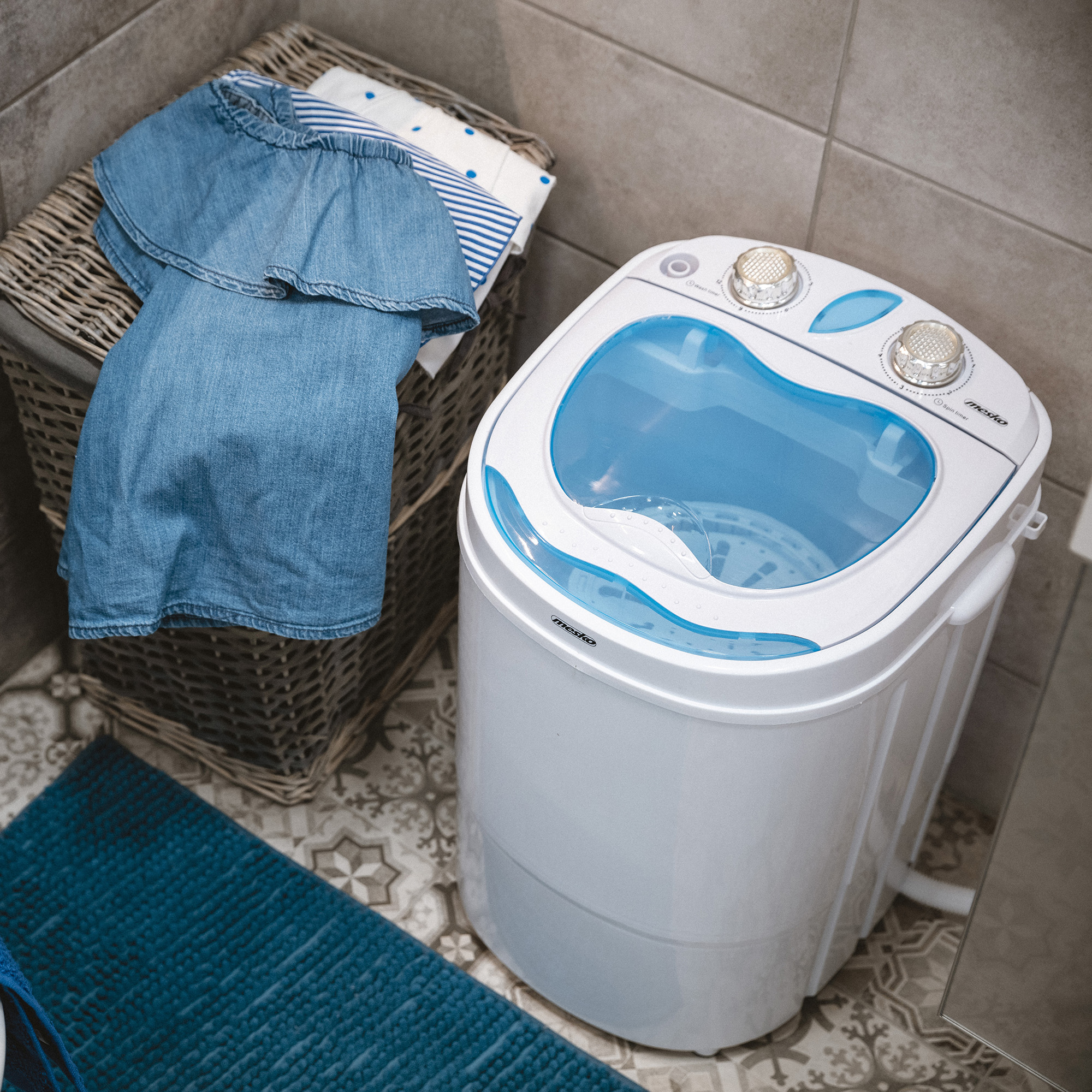 Biała pralka z niebieskimi akcentami, kosz z ubraniami i niebieska mata łazienkowa na wzorzystych kafelkach.