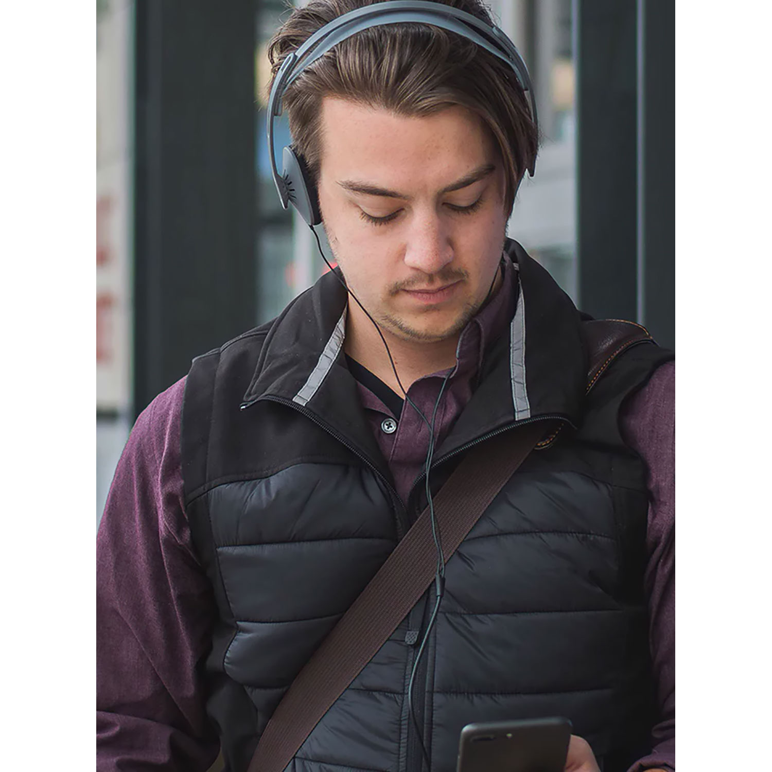 Man met koptelefoon, kijkt naar zijn telefoon, draagt een zwart vest.