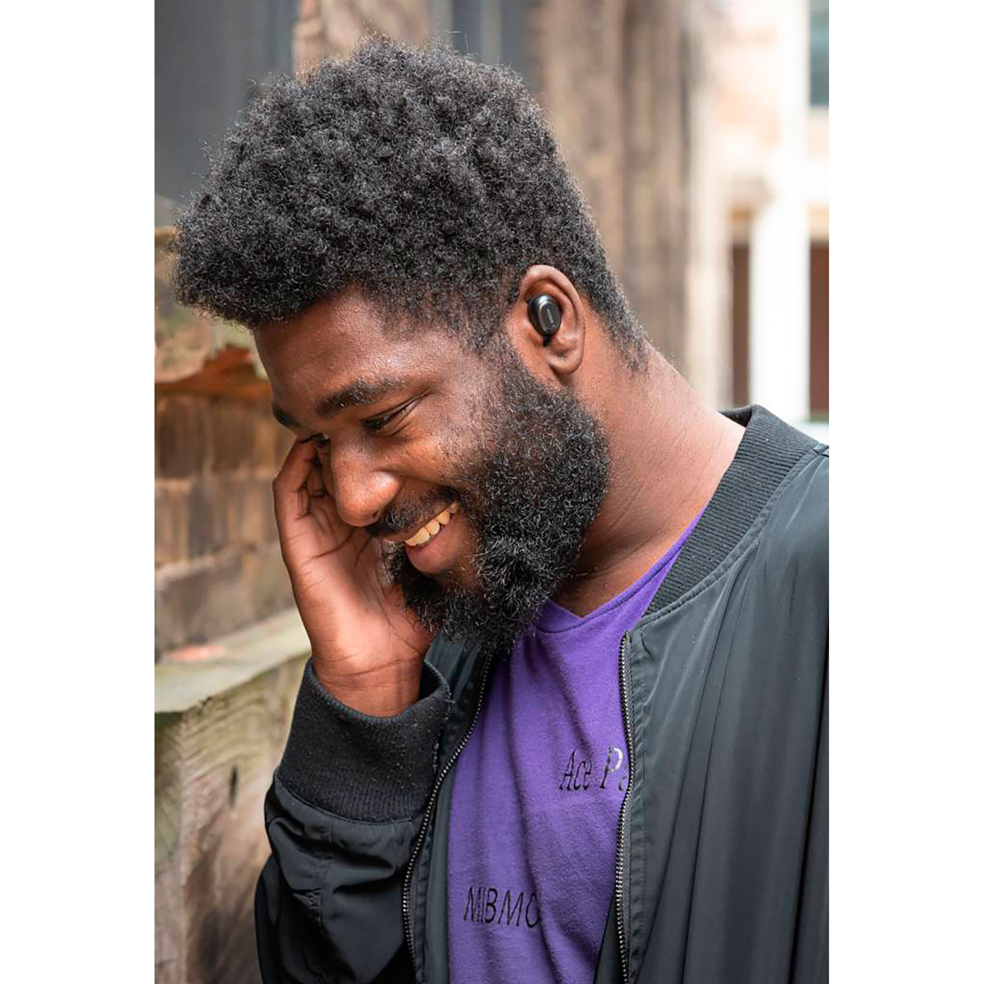 Un uomo sorridente con la barba che indossa auricolari, in un ambiente esterno.
