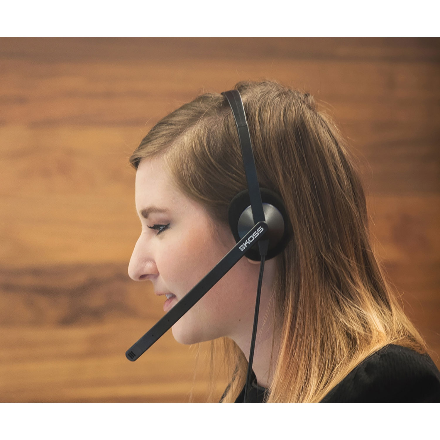 Kobieta nosząca czarny zestaw słuchawkowy Koss z mikrofonem, w profilu, uśmiechnięta. Jej włosy są długie i jasnobrązowe.