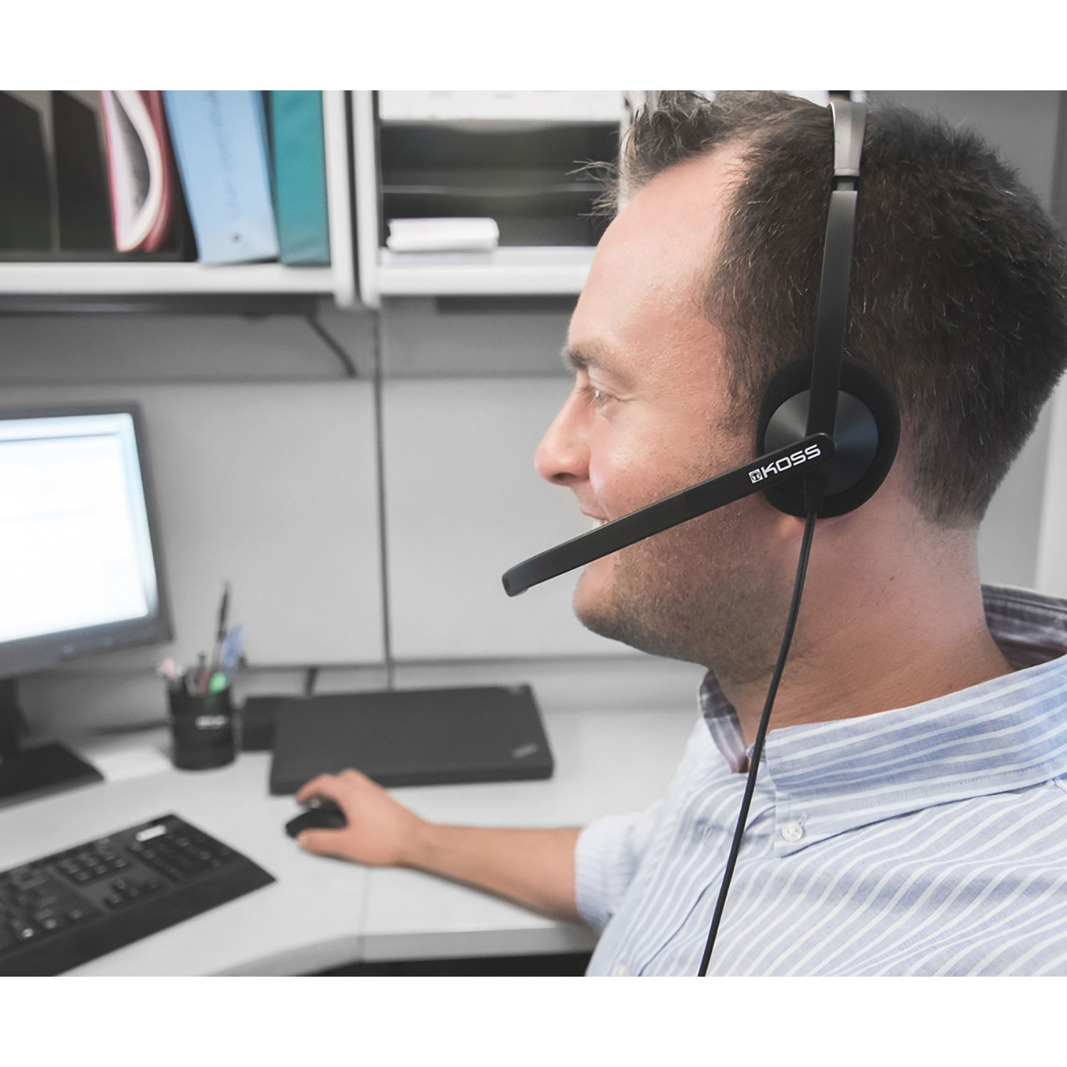 Mężczyzna w słuchawkach, uśmiechnięty, pracuje przy biurku z monitorem i klawiaturą.