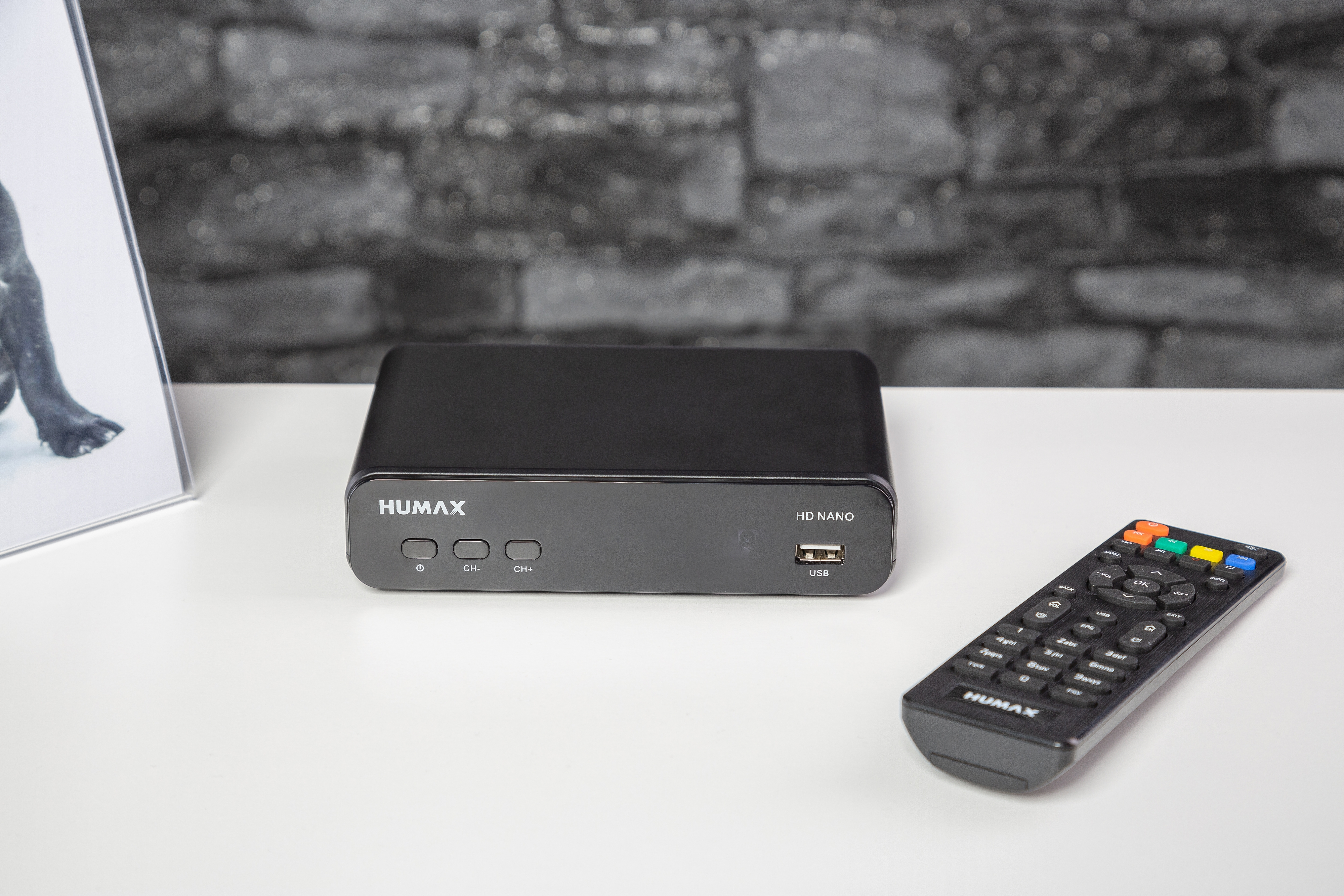 Eine schwarze Humax HD Nano Set-Top-Box und Fernbedienung. Die Box steht auf einer weißen Oberfläche mit einer Backsteinmauer.