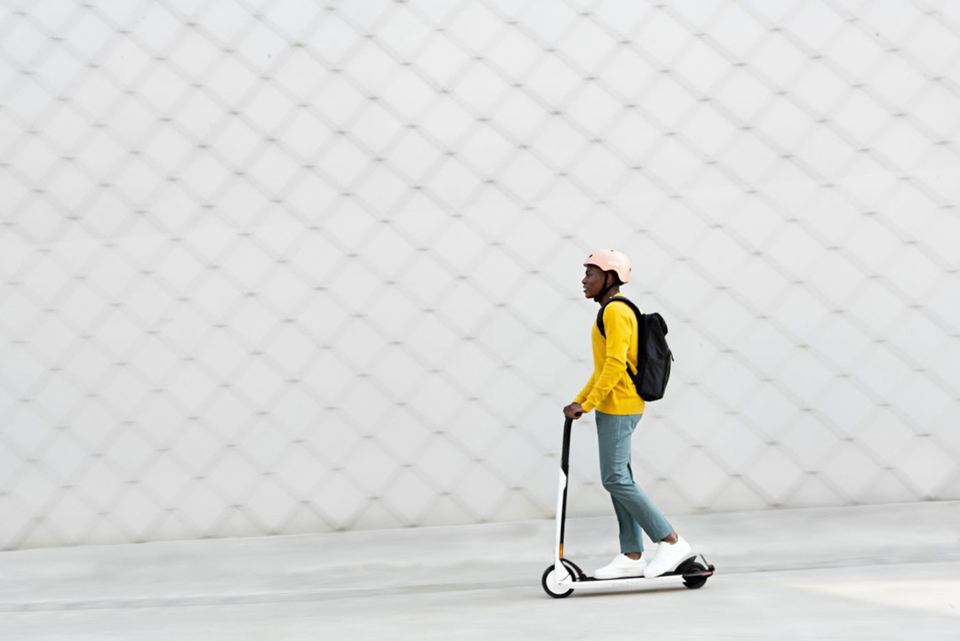 Mann auf einem Roller. Trägt gelbes Hemd, Helm und Rucksack. Fährt auf Betonfläche nahe einer weißen Wand.