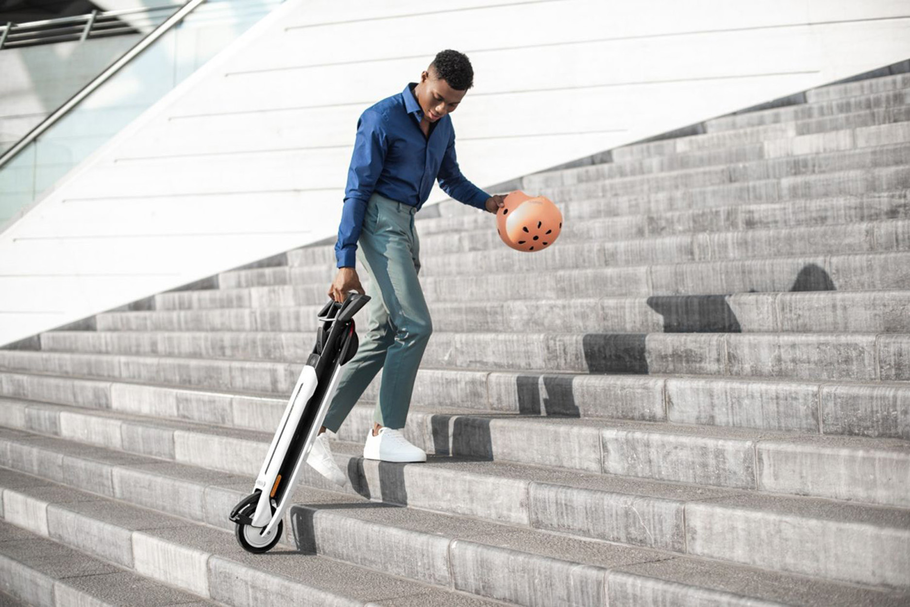 Mann geht Treppe runter, trägt zusammengeklappten Roller und Helm. Blaues Hemd, graue Hose, weiße Schuhe.