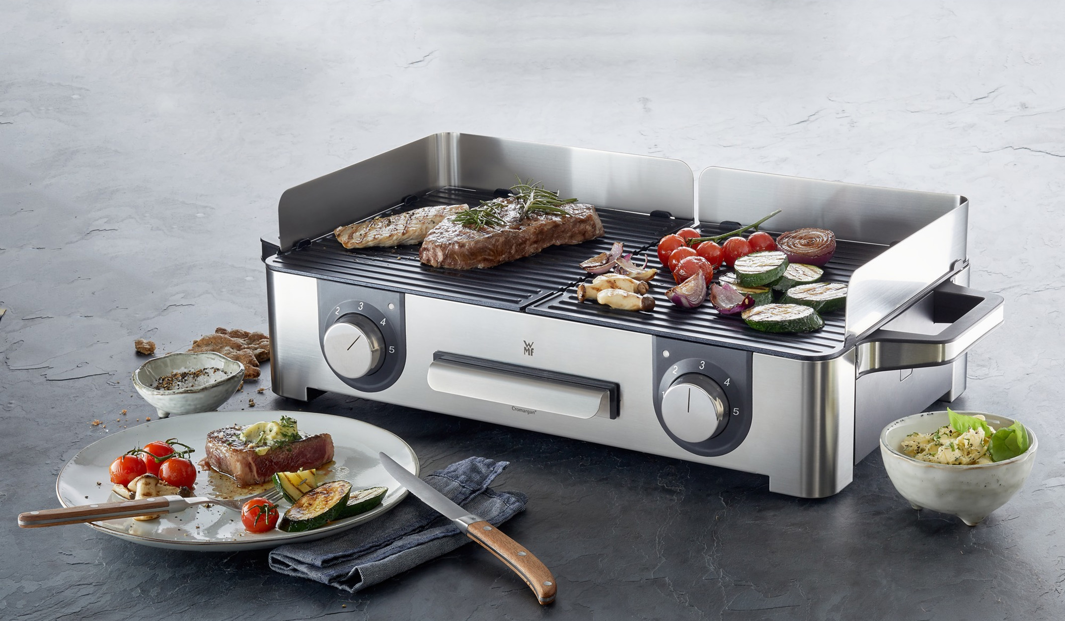 Grill wewnętrzny ze stali nierdzewnej z grillowanym stekiem, warzywami i dodatkami na stole.