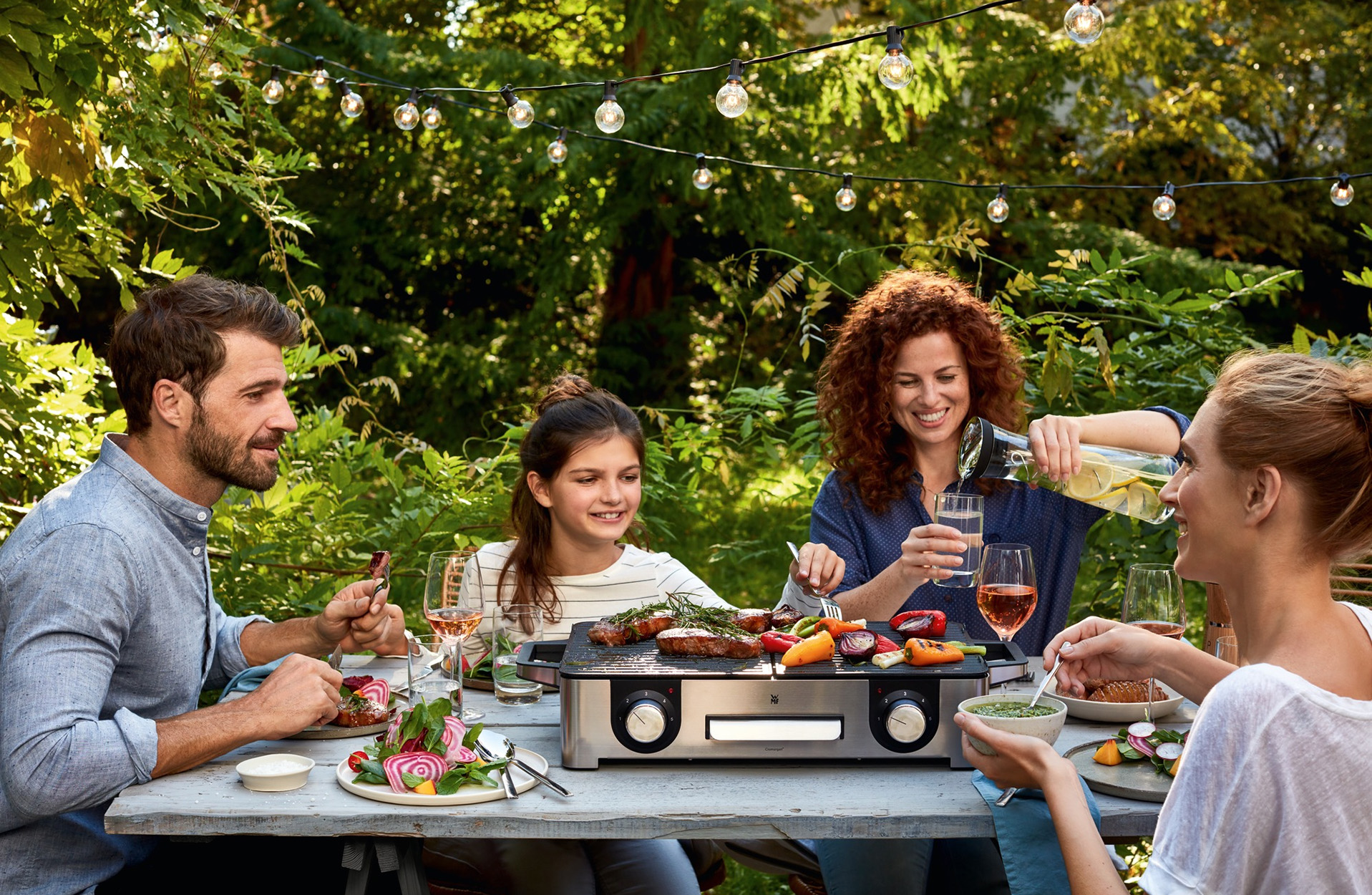 Rodzina grilluje na świeżym powietrzu. Ludzie wokół grilla jedzą i piją. Zielone drzewa w tle.
