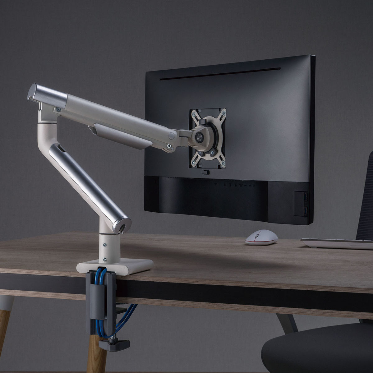 Een monitorarm die een computermonitor op een bureau houdt. Een muis en een laptop zijn zichtbaar.