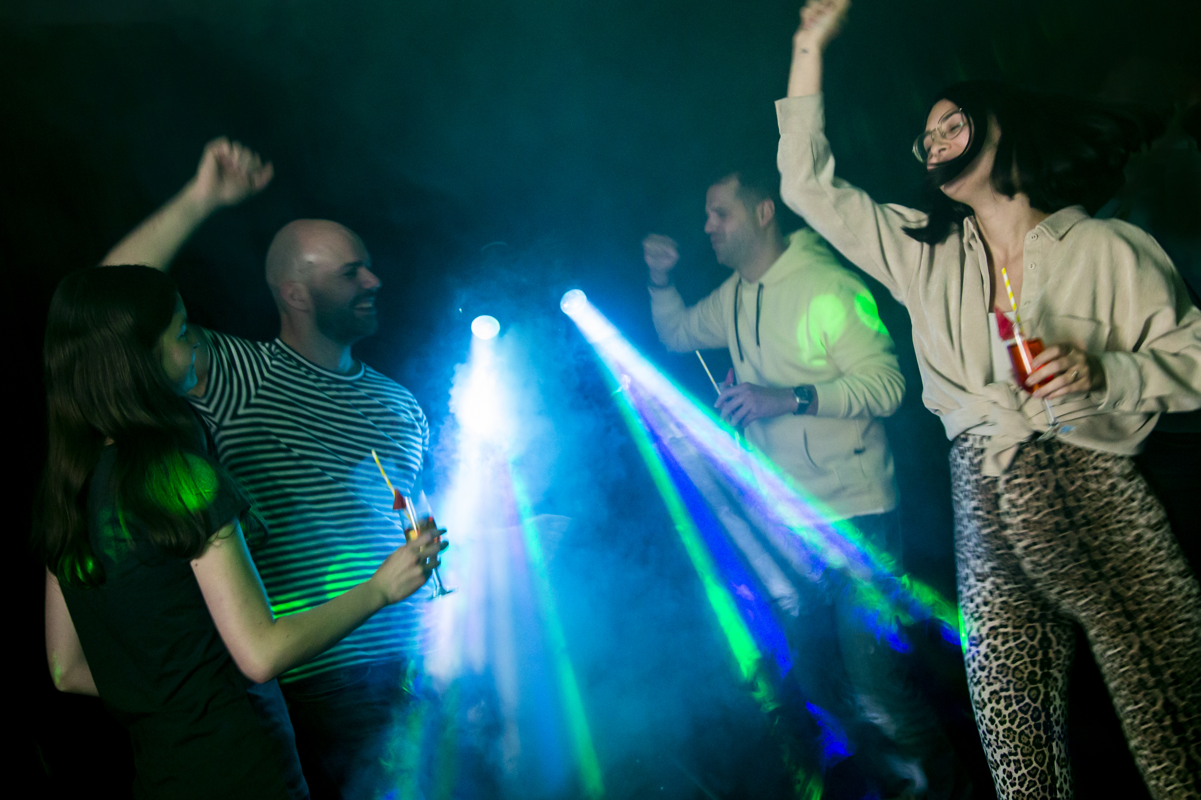 Tanzende Menschen auf einer Party mit blauem und grünem Licht, Getränken und dunklem Hintergrund.