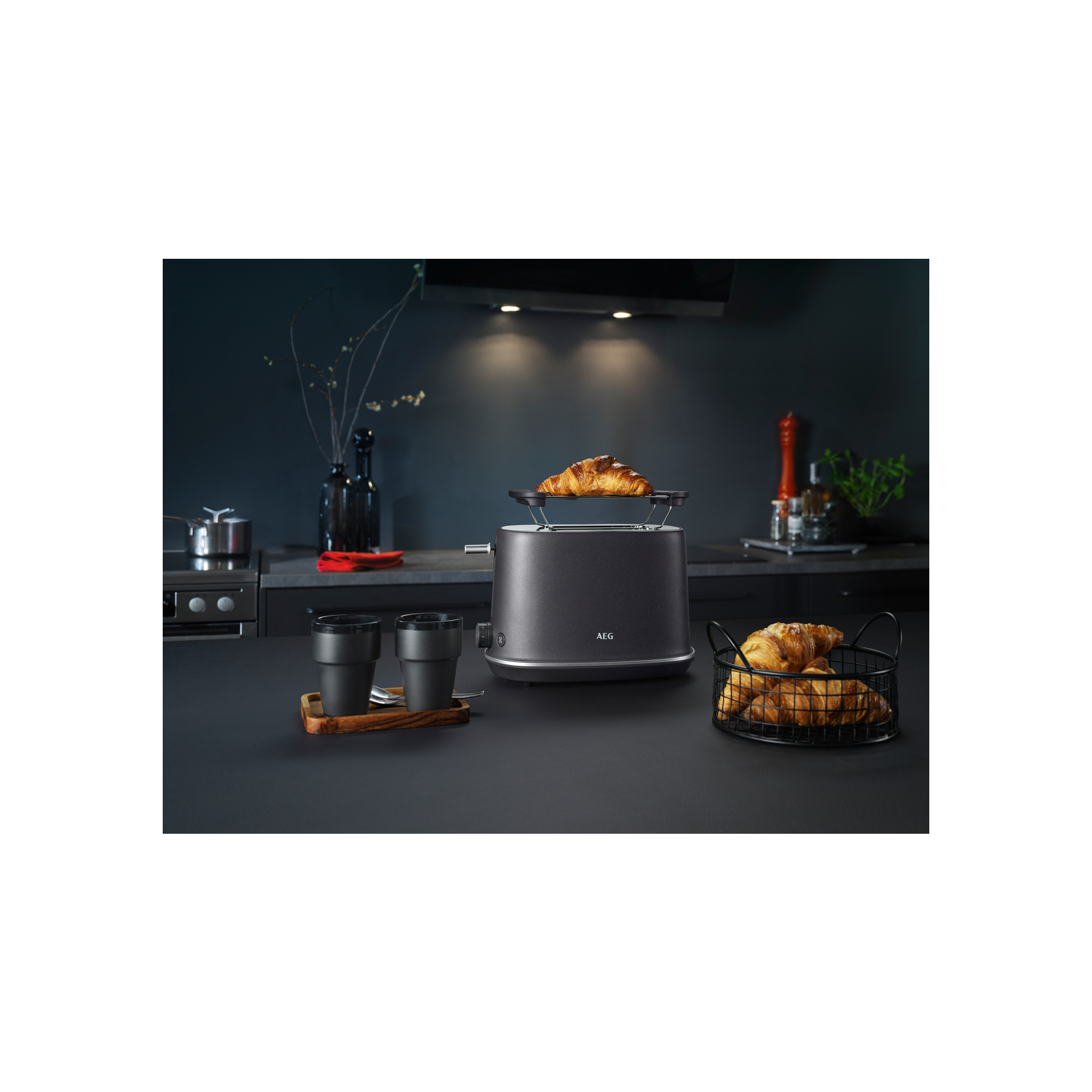 Eine Küchenszene mit einem Toaster, der Croissants toastet. Zwei Tassen und ein Korb mit Croissants stehen auf der Theke.