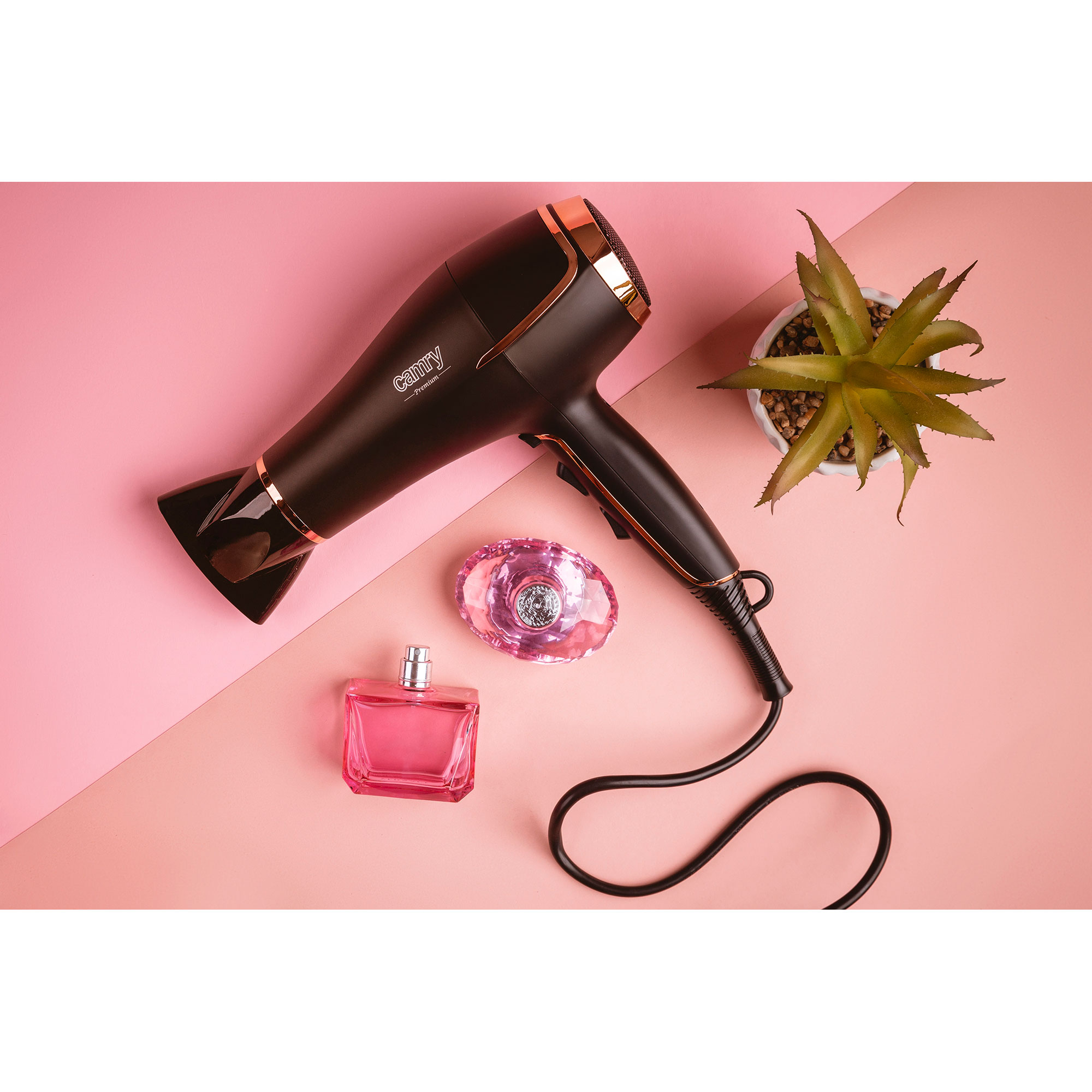 Suszarka do włosów, perfumy i roślina na różowej powierzchni. Czarna suszarka ze złotymi akcentami.