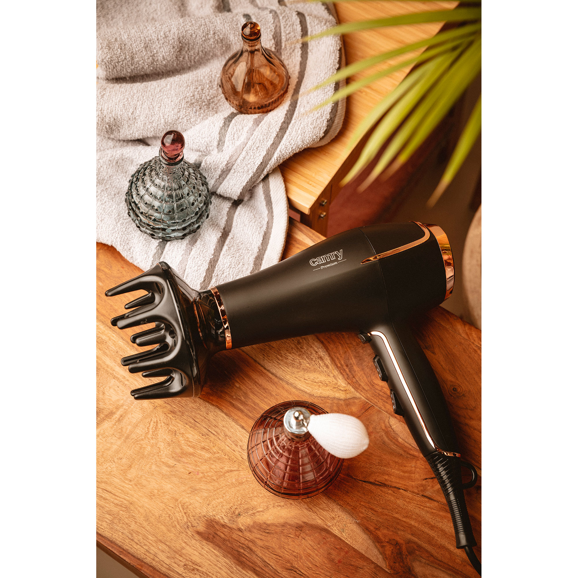 Susząca do włosów i butelki perfum na drewnianym stole z ręcznikiem i rośliną w tle.