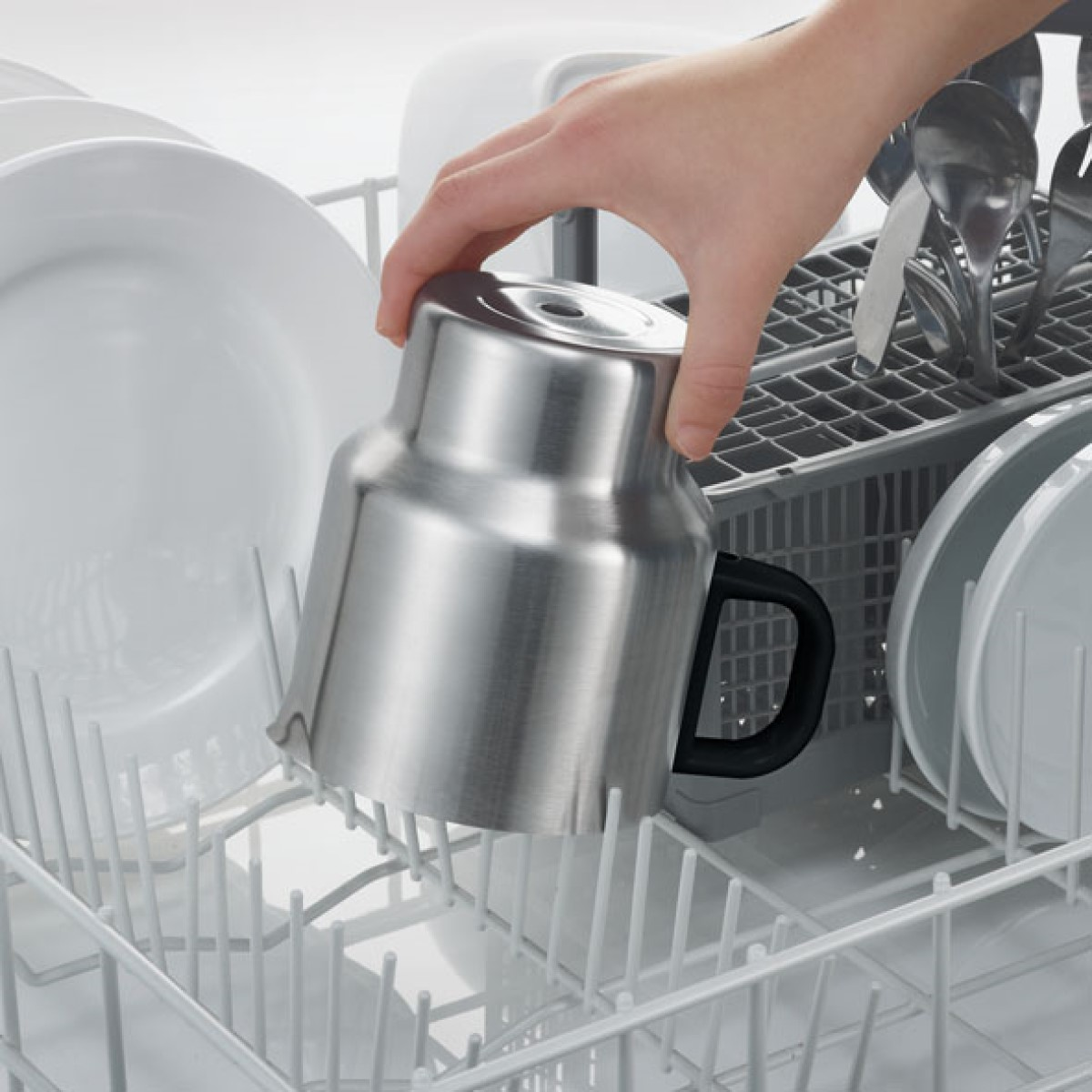 A hand places a metal mug into a dishwasher rack. Other dishes are in the background.
