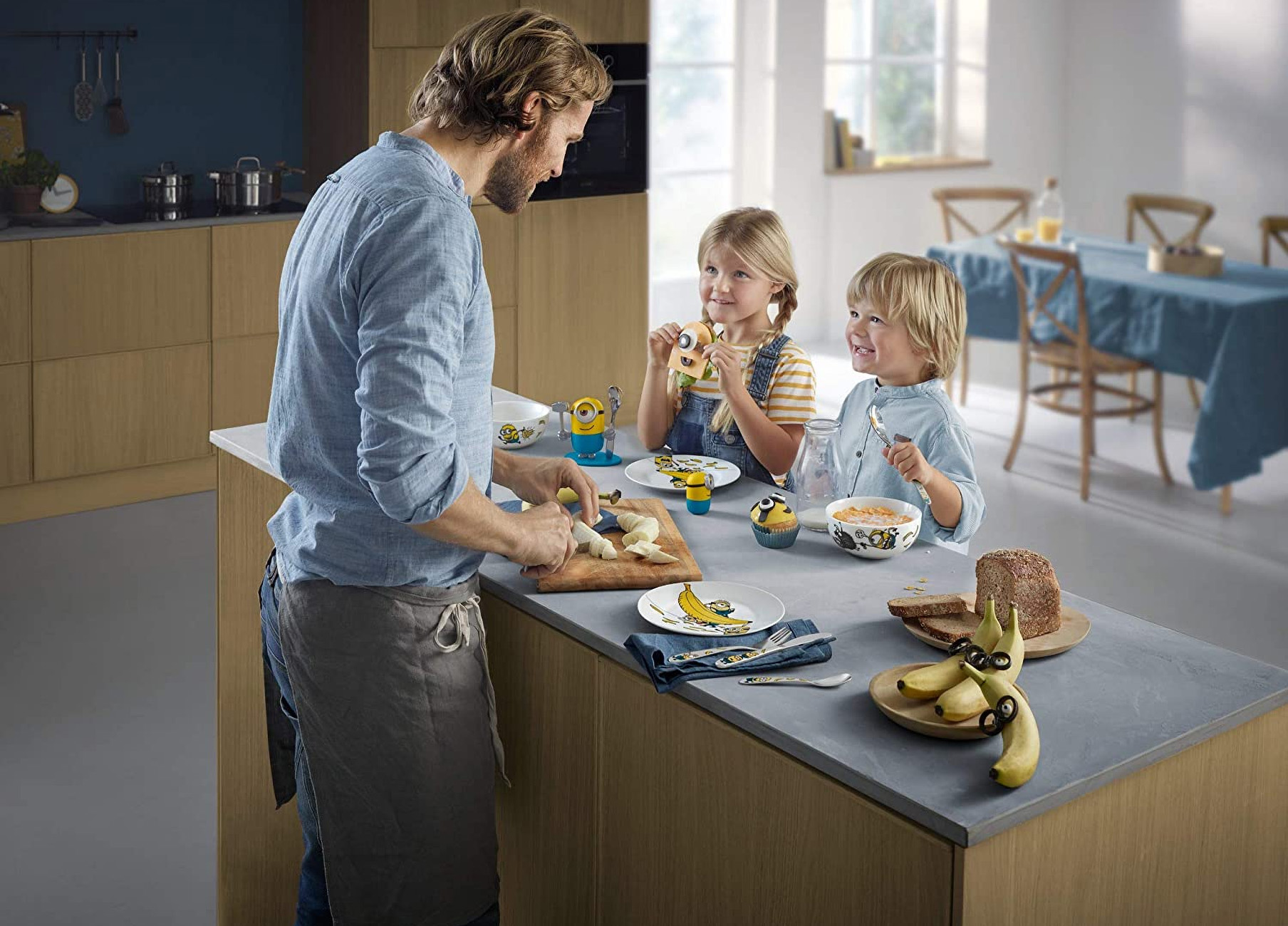 Ein Mann schneidet Bananen an einem Tisch, an dem zwei Kinder essen. Minion-Geschirr ist vorhanden.