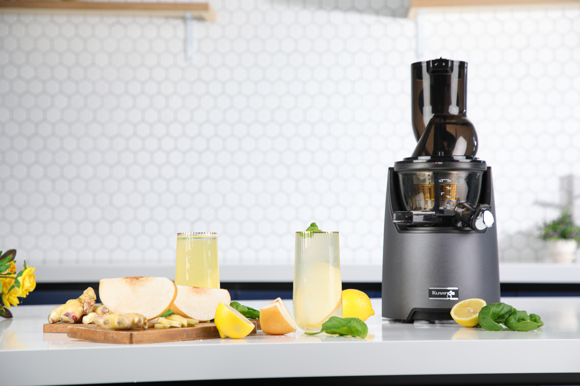 Exprimidor con dos bebidas, rodajas de fruta e ingredientes sobre una mesa blanca en una cocina.
