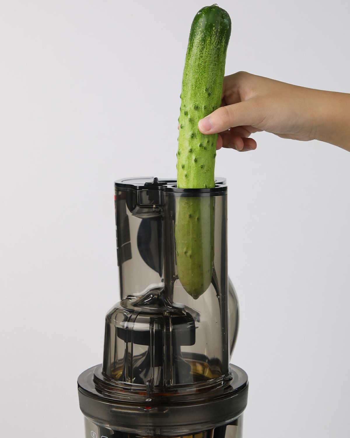 Una persona inserta un pepino verde en una licuadora oscura. El fondo es blanco.