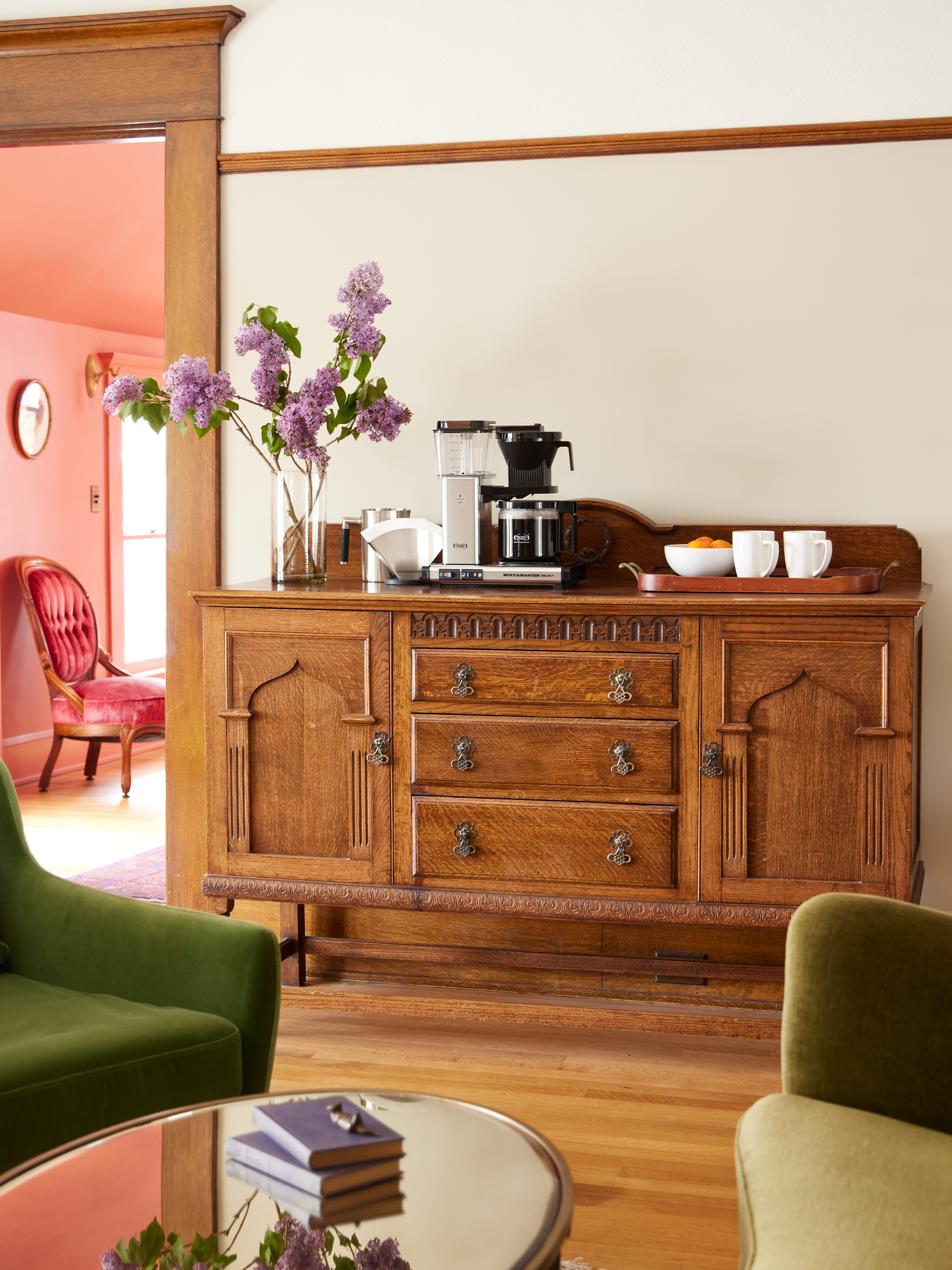 Drewniany kredens z ekspresem do kawy i kwiatami. Ekspres do kawy i kubki na wierzchu. Widoczny czerwony pokój.