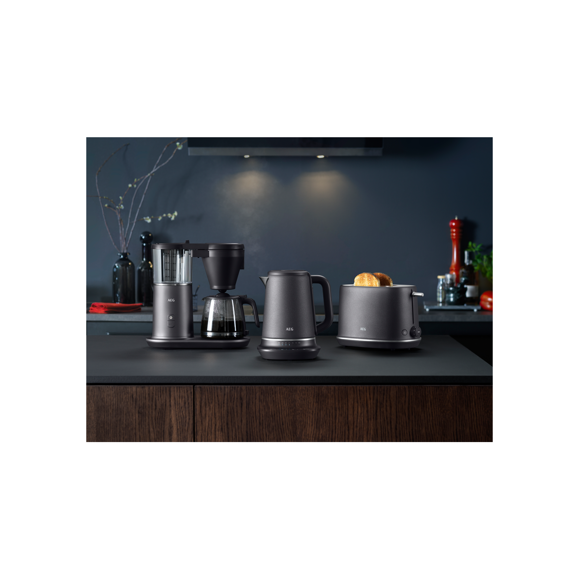 Mroczna scena kuchenna z urządzeniami AEG: ekspres do kawy, czajnik, toster i taca. Czarne blaty.