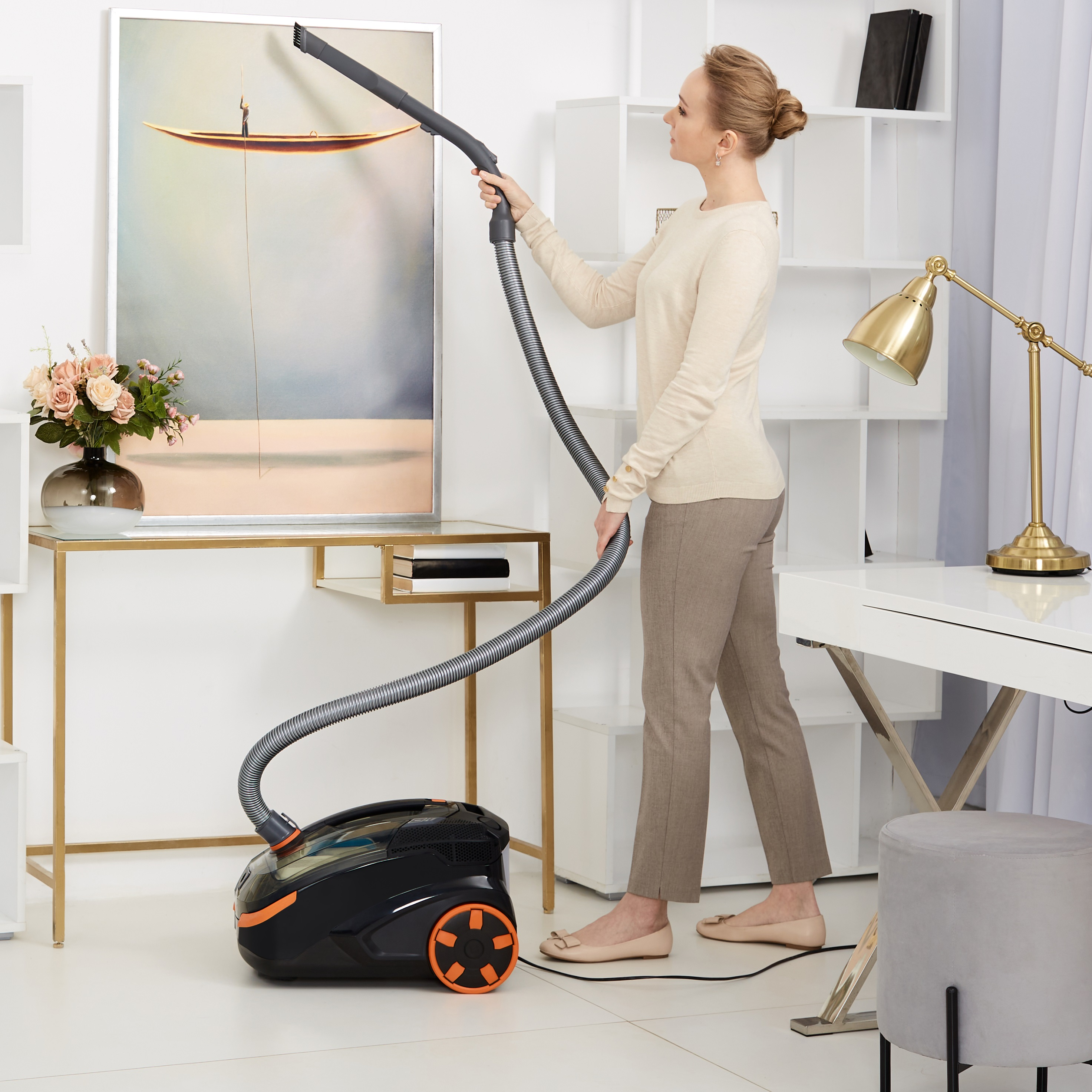 Frau, die ein Bild an einer Wand mit einem schwarz-orangenen Staubsauger auf weißem Hintergrund absaugt.