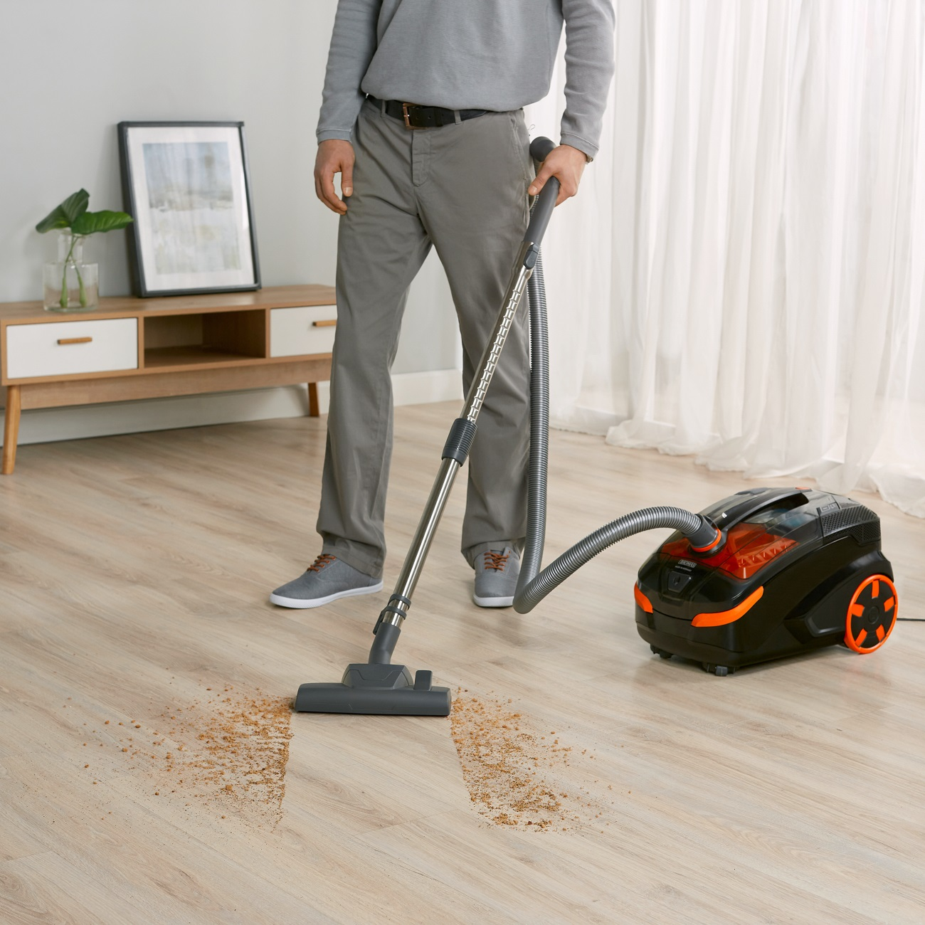 Ein Mann saugt einen Holzboden. Ein Staubsauger mit orangefarbenen Akzenten befindet sich rechts.