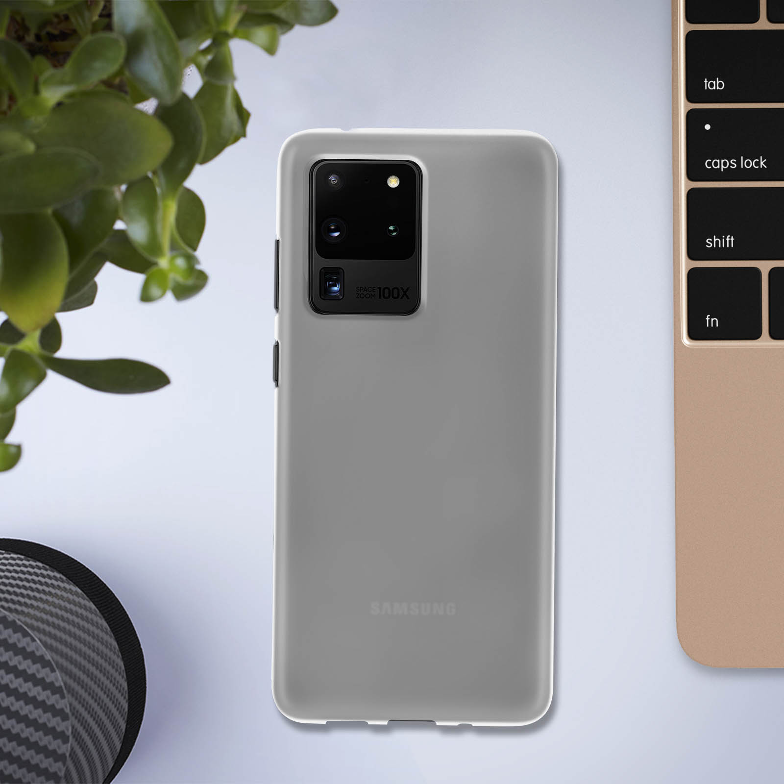 Ein graues Samsung-Telefon in einer grauen Hülle liegt auf einer weißen Oberfläche. Eine grüne Pflanze und eine Tastatur sind sichtbar.