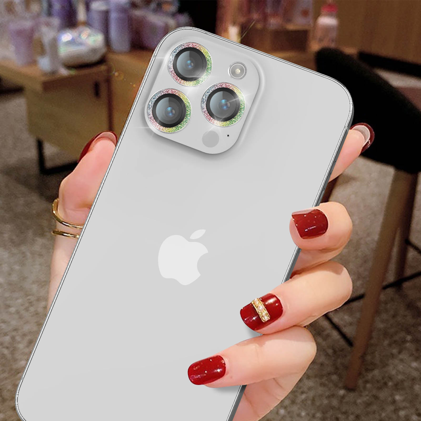 La main d'une personne tenant un téléphone argenté avec des protections d'objectif, des ongles rouges et une bague dorée.