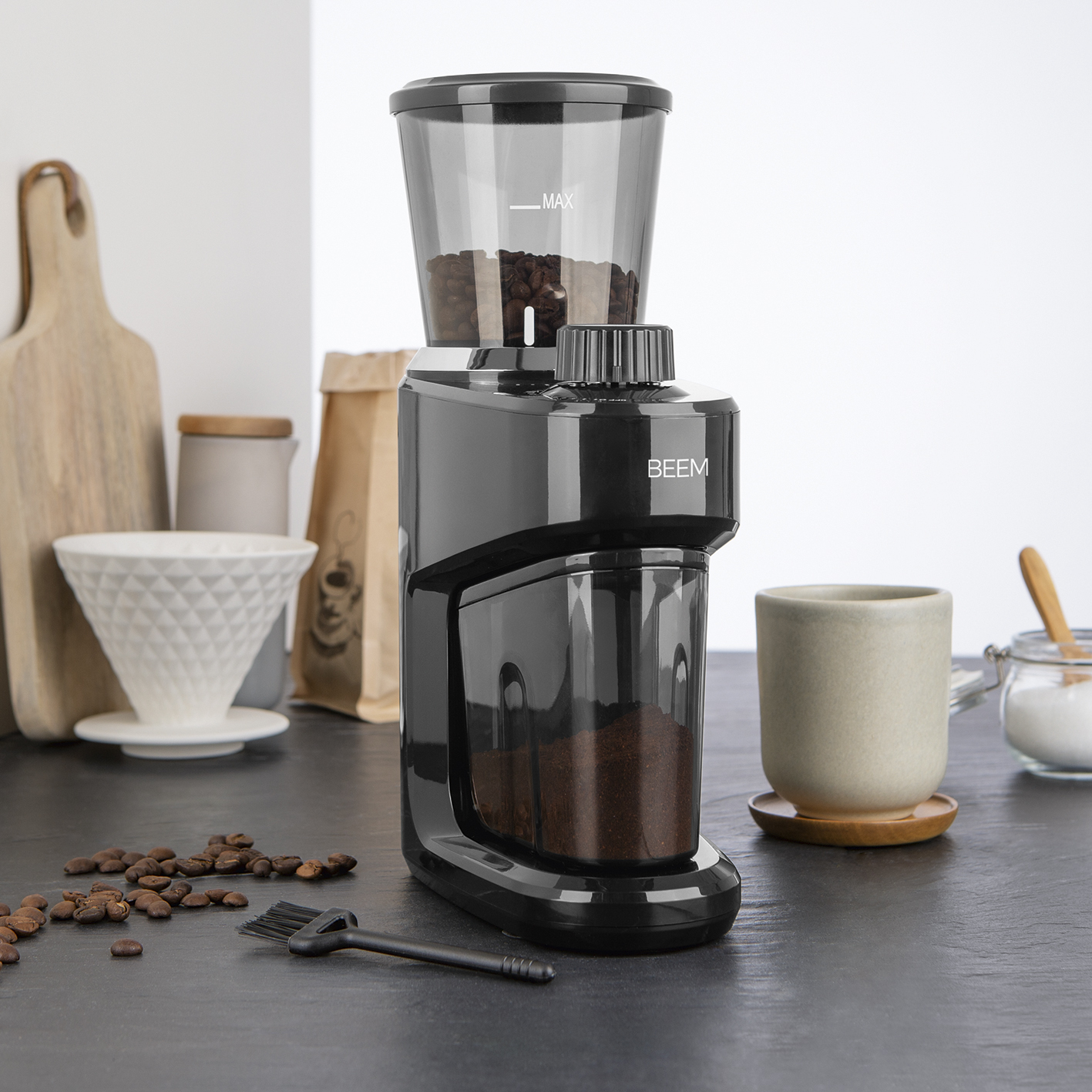Schwarze Kaffeemühle mit Bohnen oben, gemahlenem Kaffee unten. Kaffeebohnen und Bürste sind vorne.