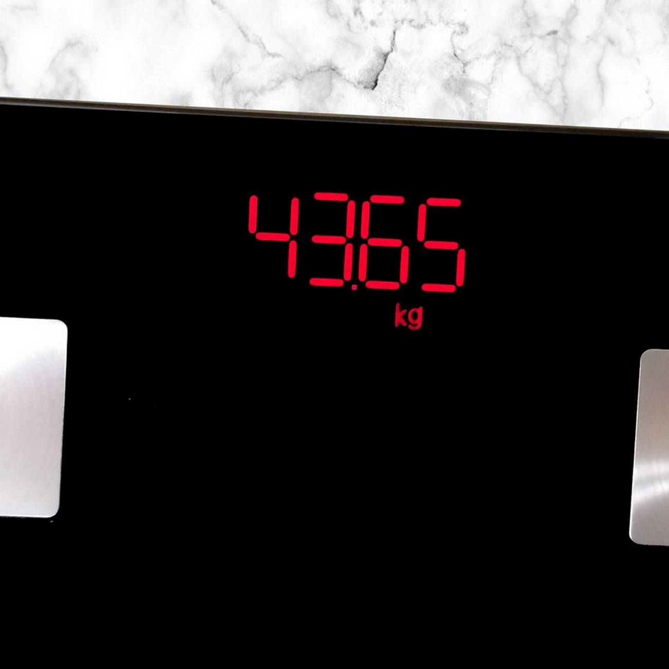 Una báscula digital negra muestra 43,65 kg en dígitos rojos. El fondo es mármol blanco.