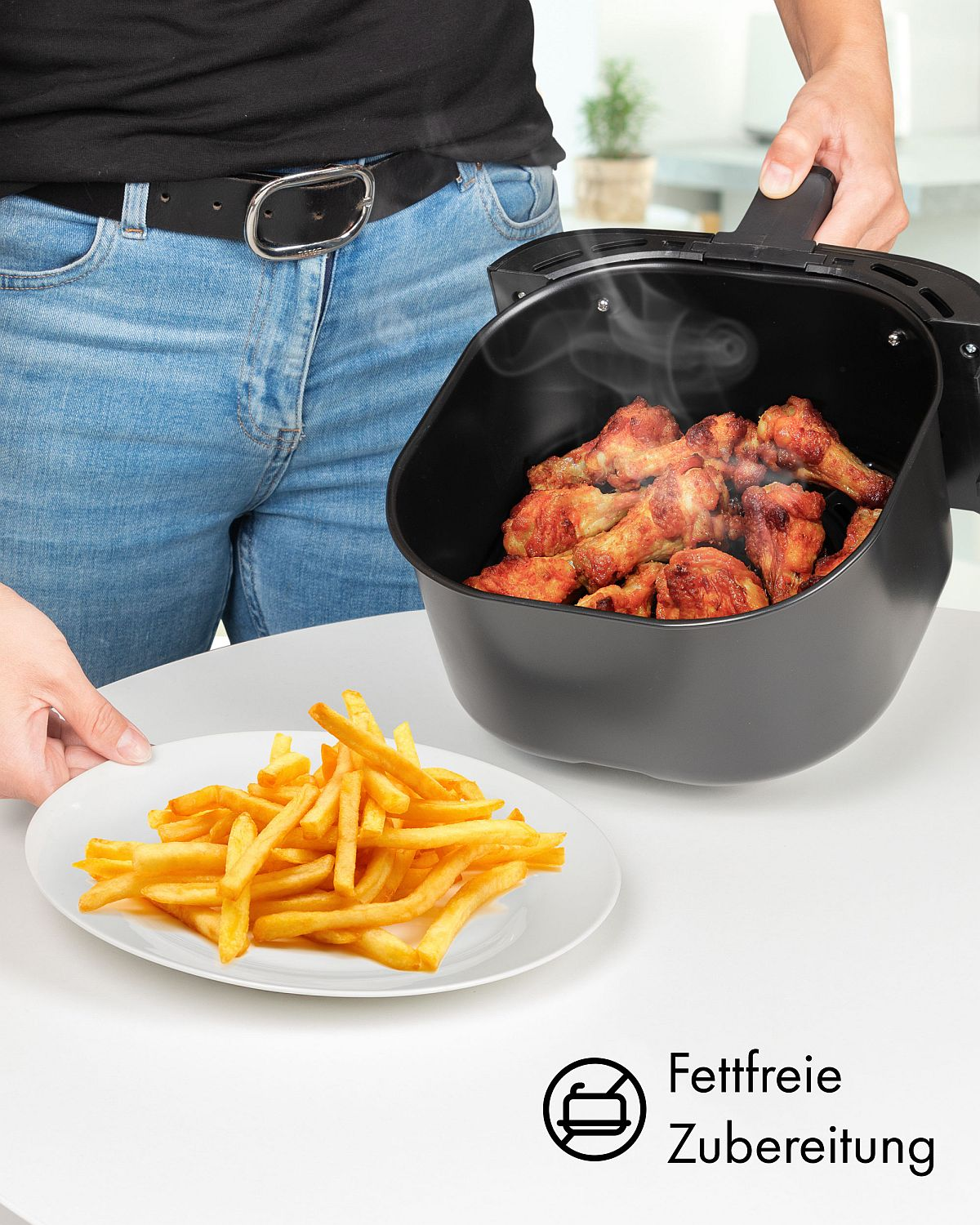 Una persona sostiene una freidora de aire con alitas de pollo cocinadas y papas fritas en un plato.
