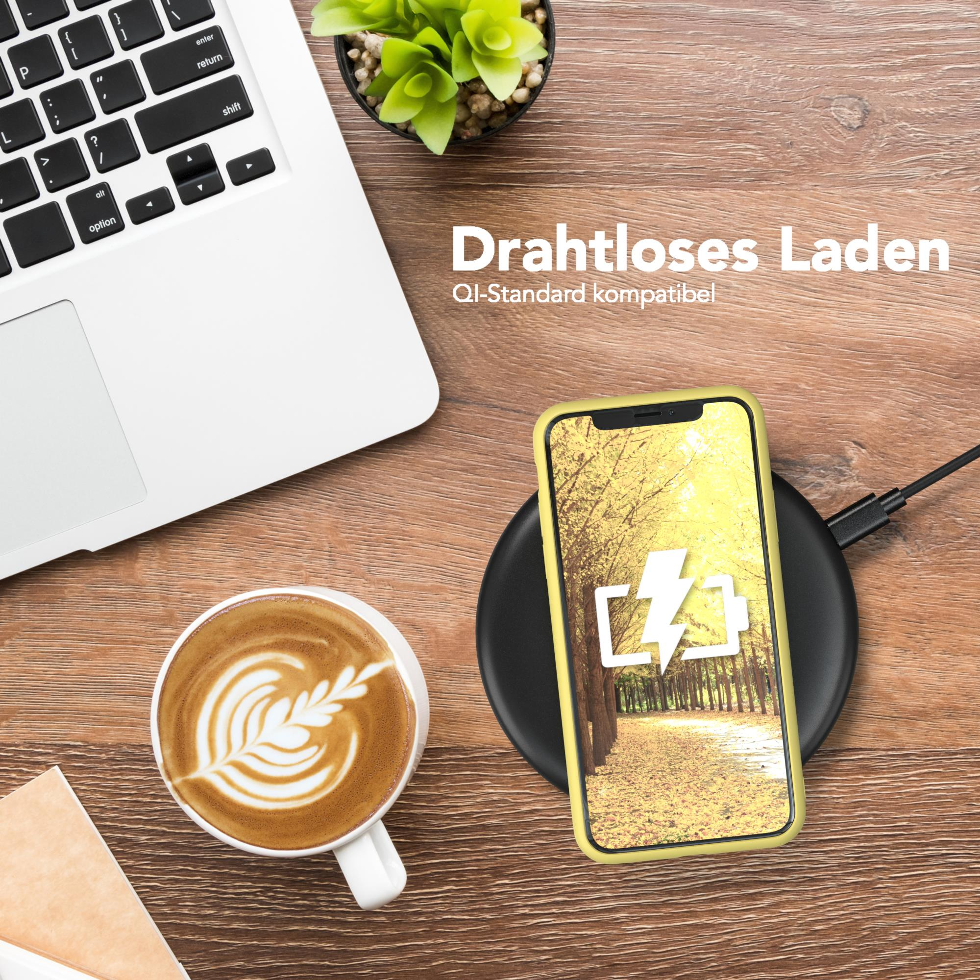 Telefon lädt auf kabellosem Ladegerät, neben Laptop und Kaffee. Holz Tisch. Eine Pflanze im Hintergrund.
