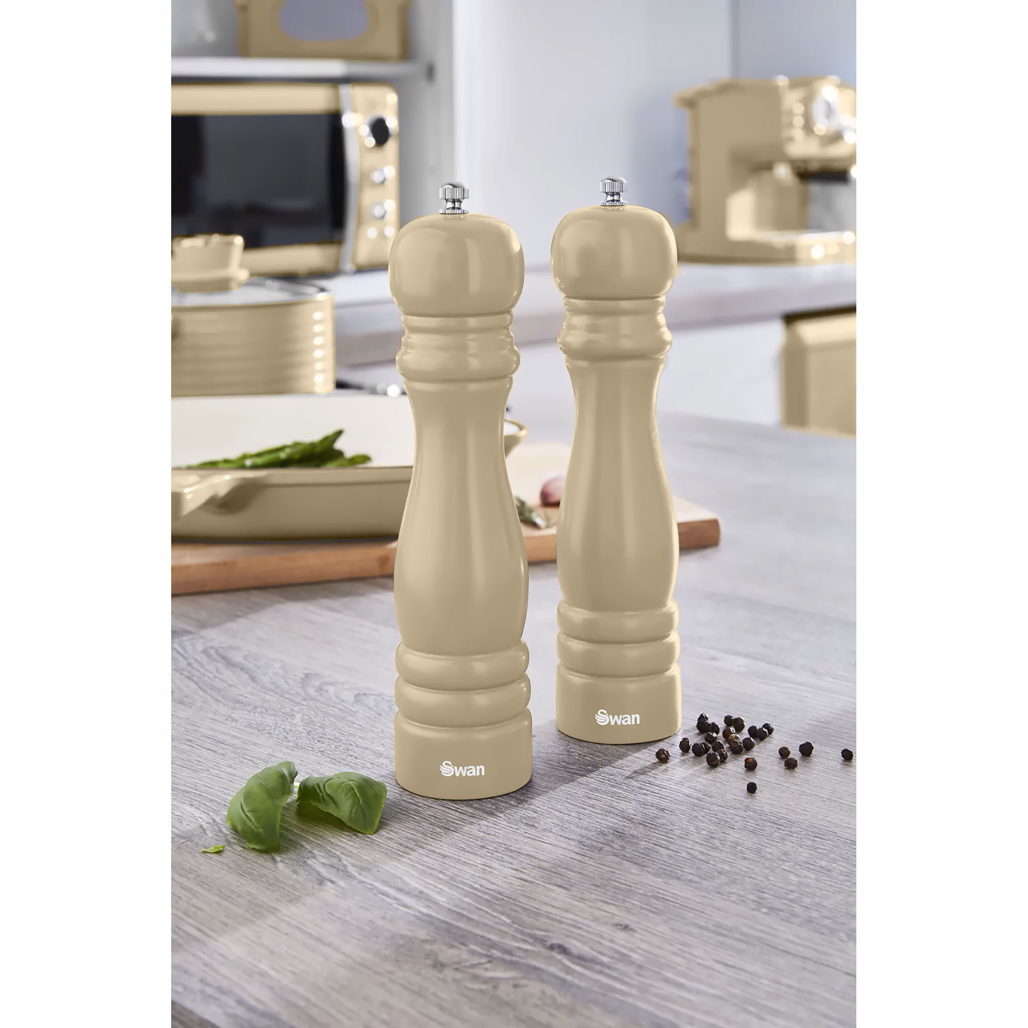 Dos molinillos de pimienta beige sobre una superficie de madera, con hojas de albahaca y granos de pimienta.