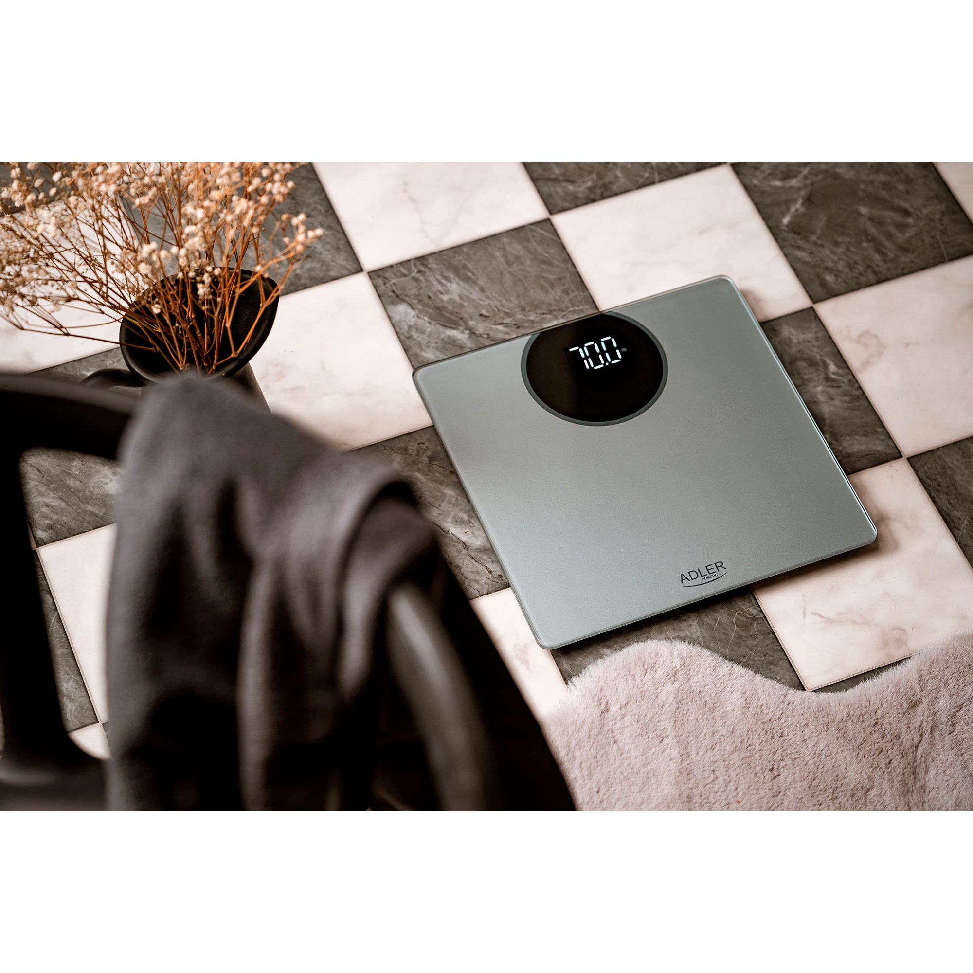 Szara cyfrowa waga łazienkowa na wzorzystej podłodze, z ręcznikiem i wazonem z kwiatami.