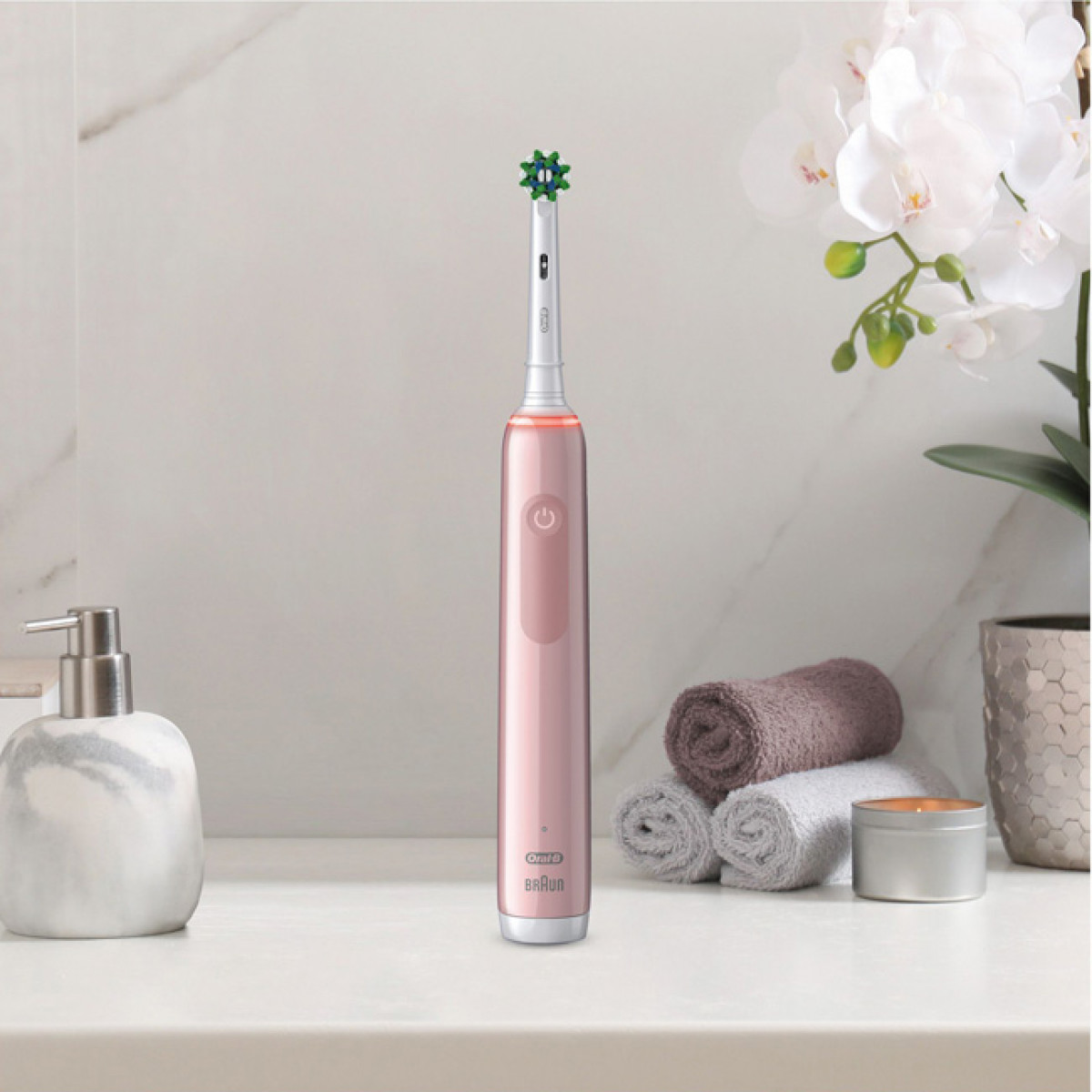 Un cepillo de dientes eléctrico rosa está sobre una superficie blanca con flores, toallas y un dispensador de jabón.