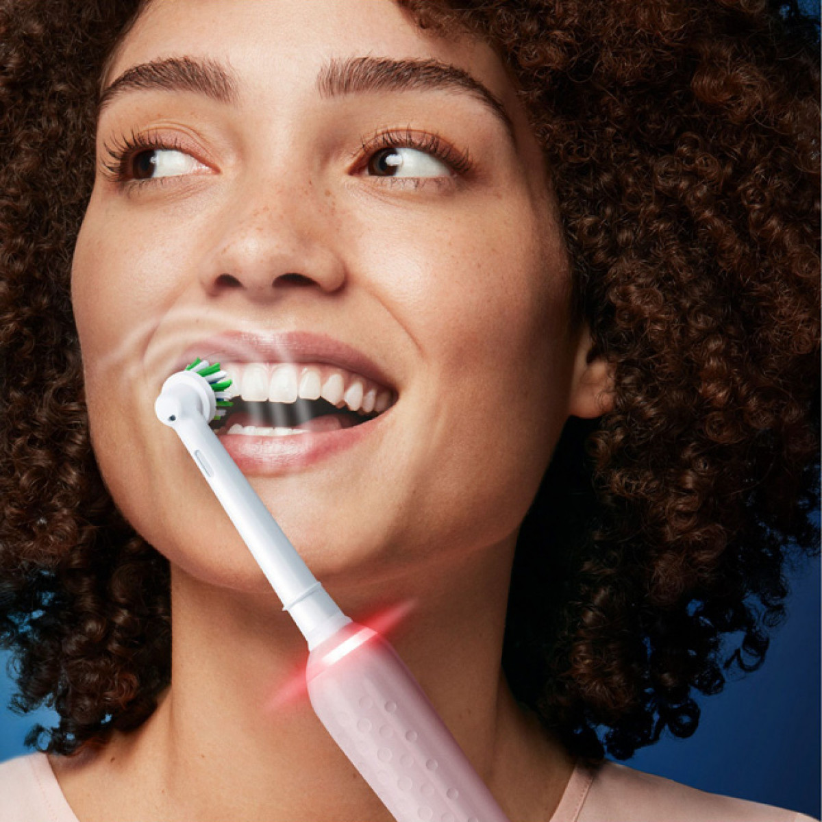 Una mujer cepillándose los dientes con un cepillo eléctrico. El cepillo de dientes es rosa y la boca está abierta.