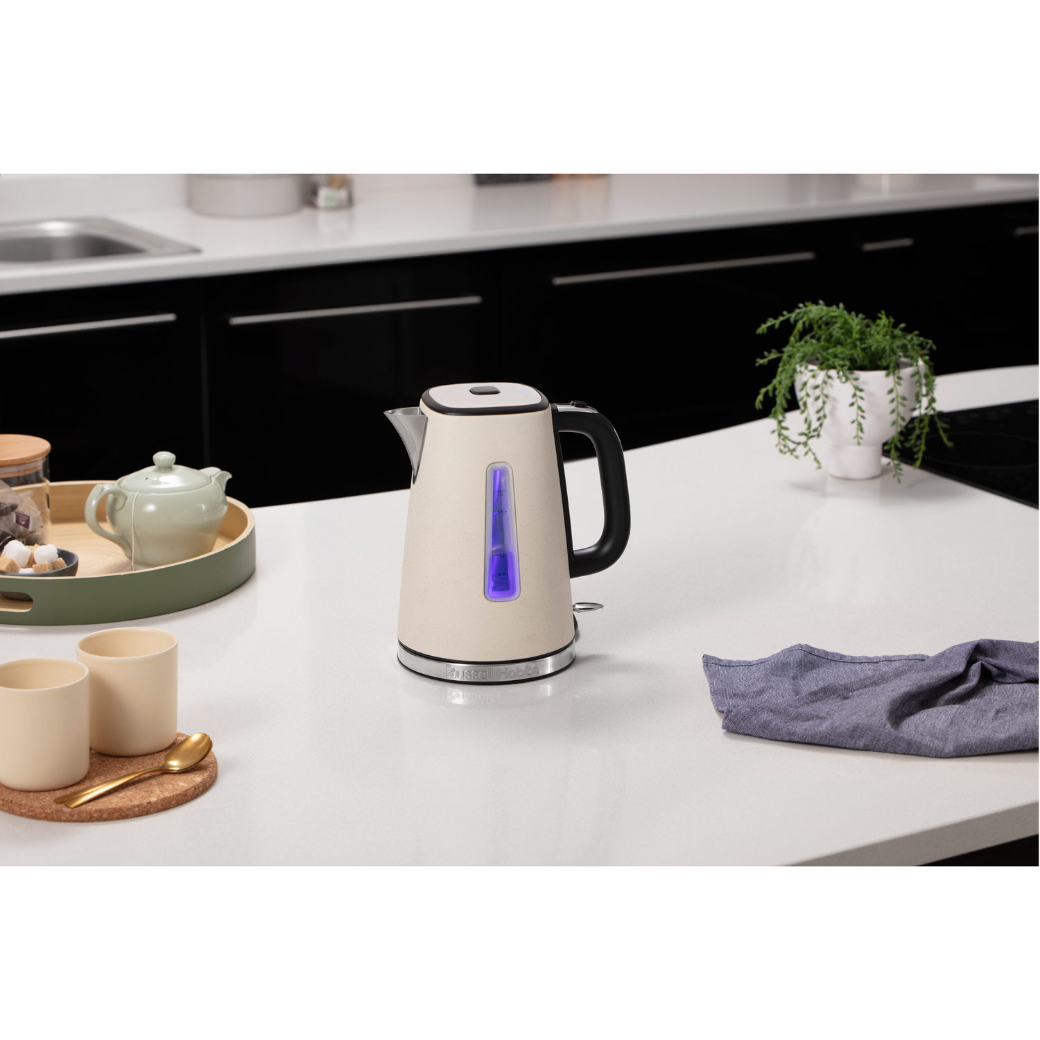 Beżowy czajnik elektryczny. Biały blat, czarne szafki i zielona roślina. Niebieskie światło. Złote akcenty.
