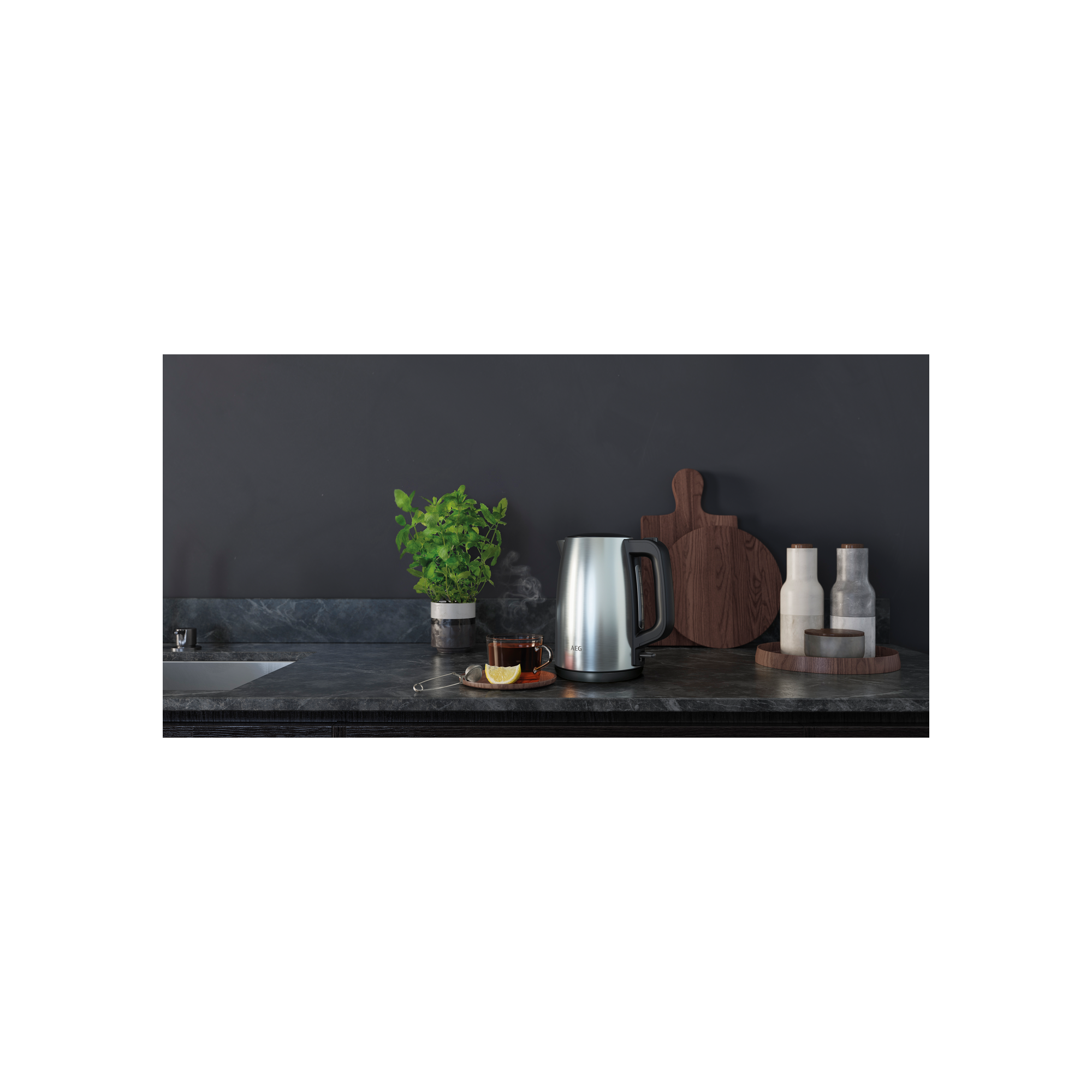 Scena cucina con bollitore argentato, tazza di tè, taglieri in legno e pianta su superficie scura.