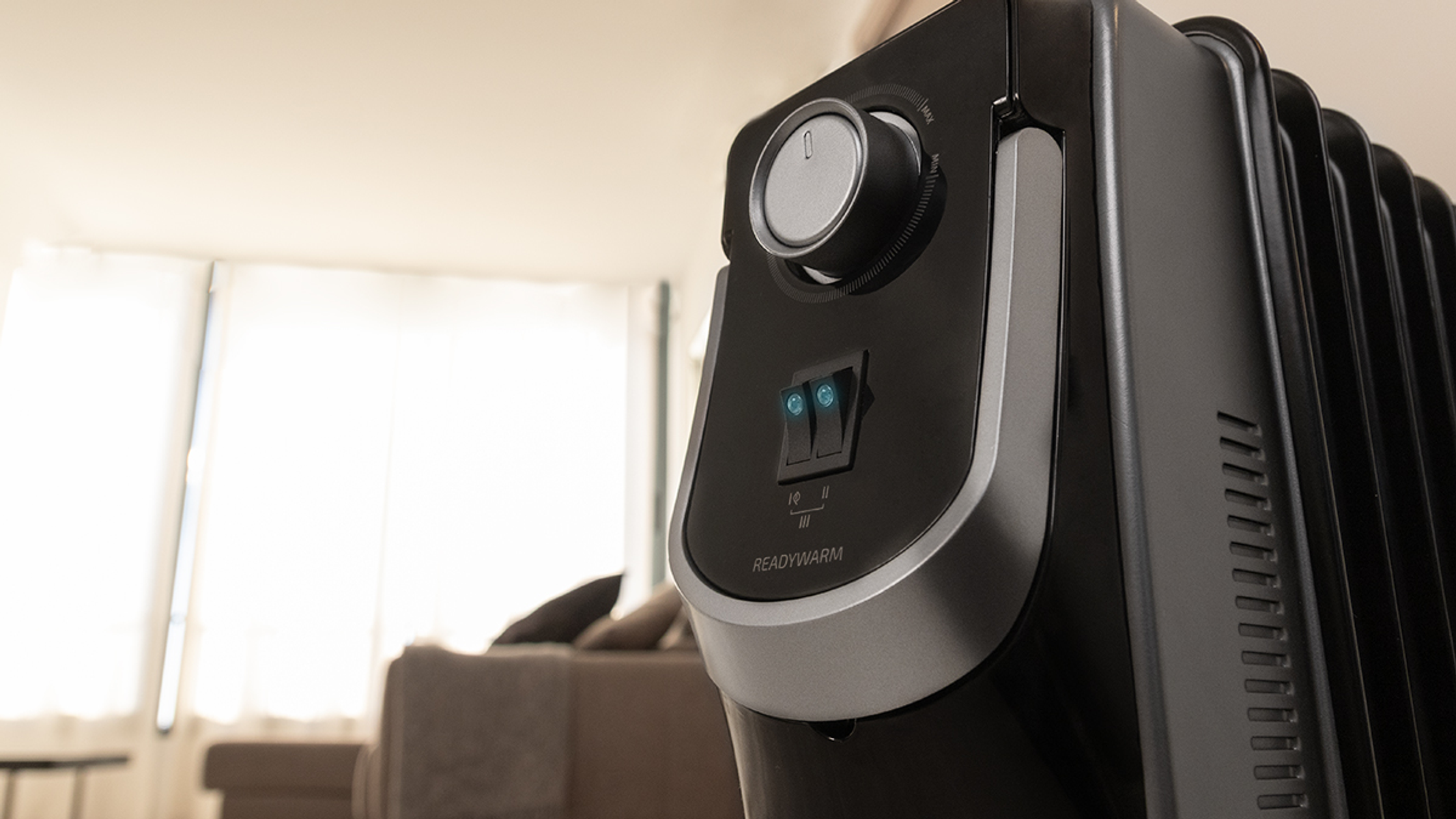 Zbliżenie czarnego grzejnika olejowego ReadyWarm z pokrętłem i przełącznikami. Rozmyty salon w tle.