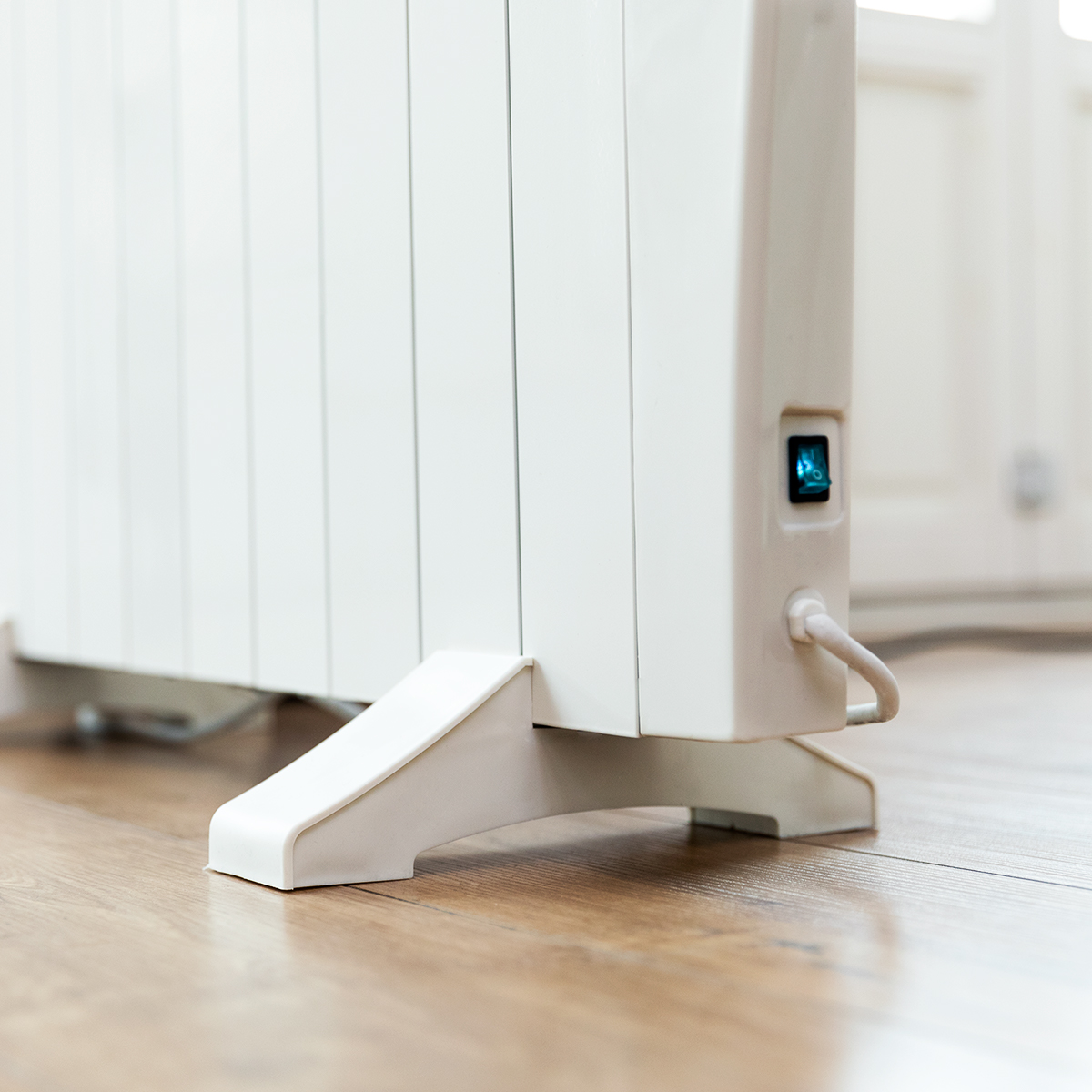Biały grzejnik elektryczny z niebieskim przyciskiem zasilania i drewnianą podłogą.