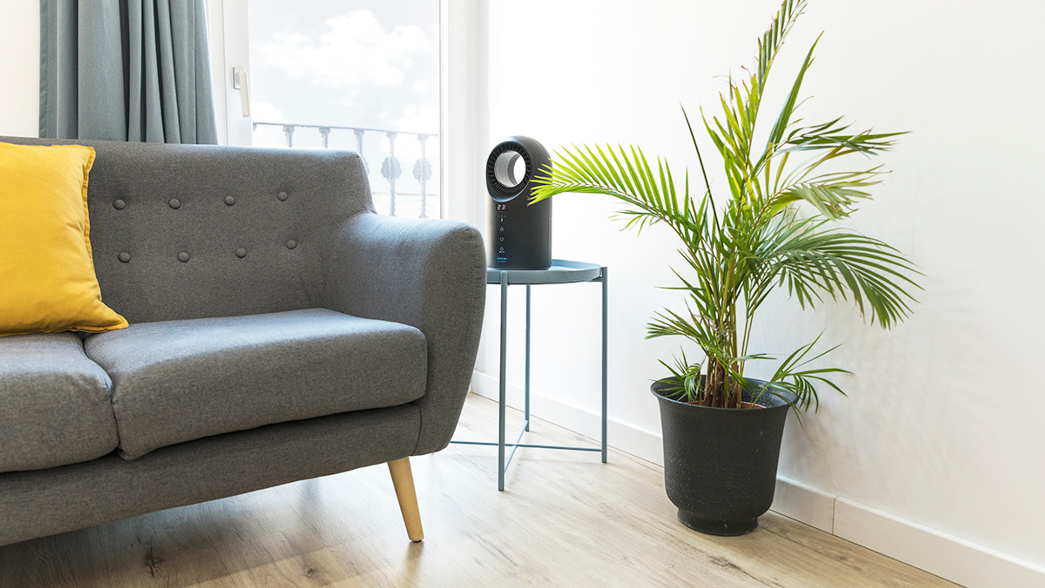 Scena w salonie z szarą sofą, żółtą poduszką, rośliną i czarnym oczyszczaczem powietrza.