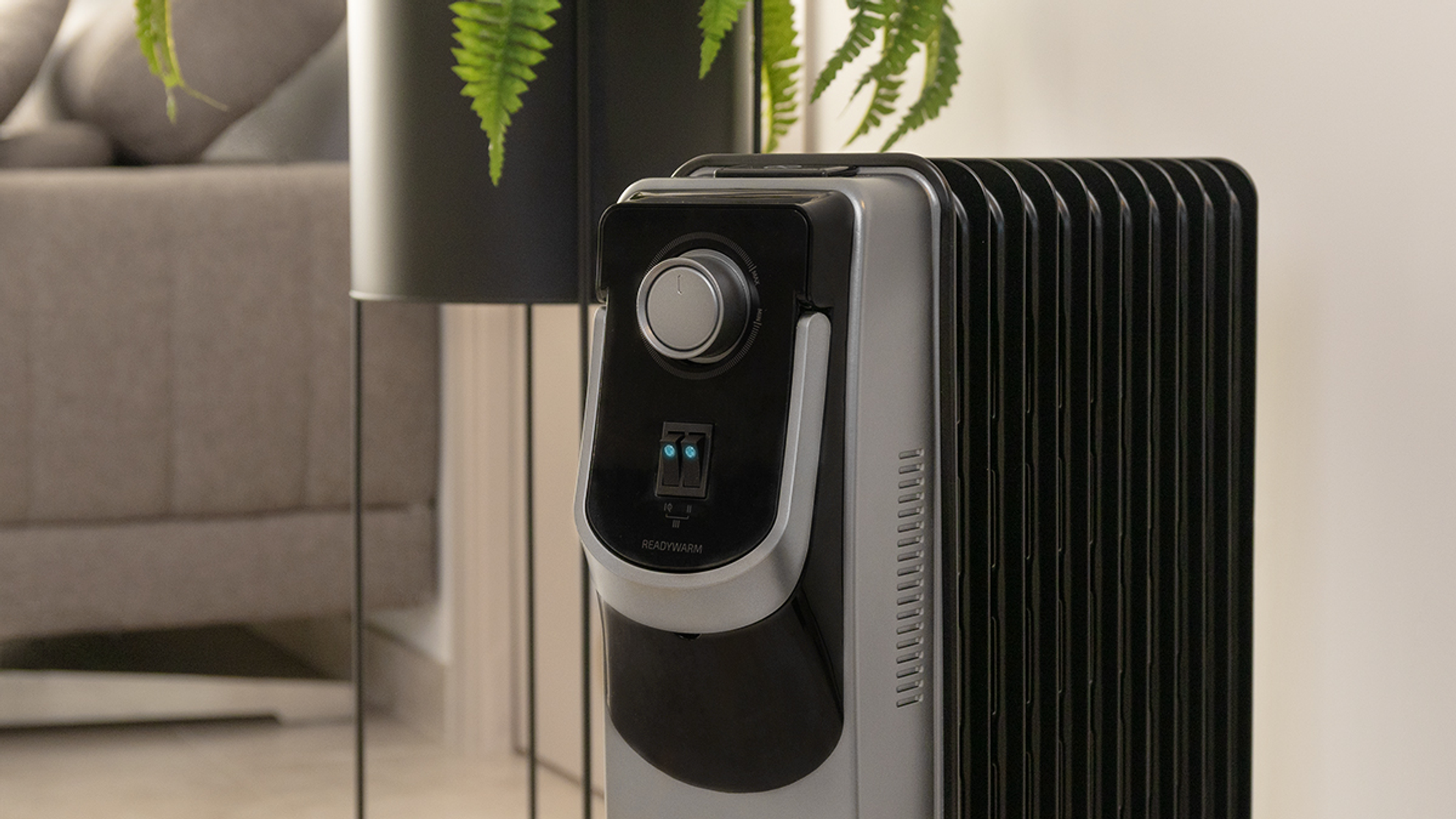 Grzejnik olejowy, czarno-srebrny, z rośliną i sofą w tle.