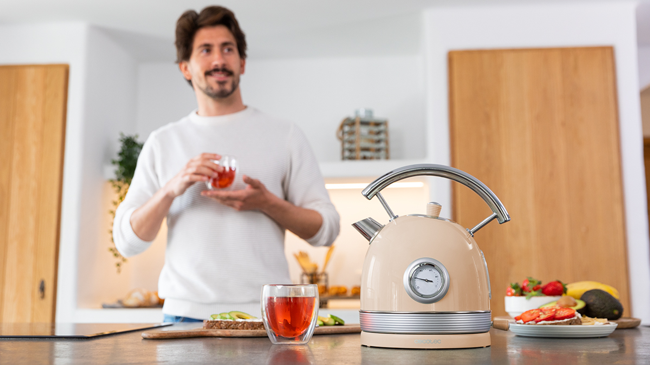 Mężczyzna w kuchni z herbatą. Czajnik i jedzenie na blacie. Jasne drewniane szafki.